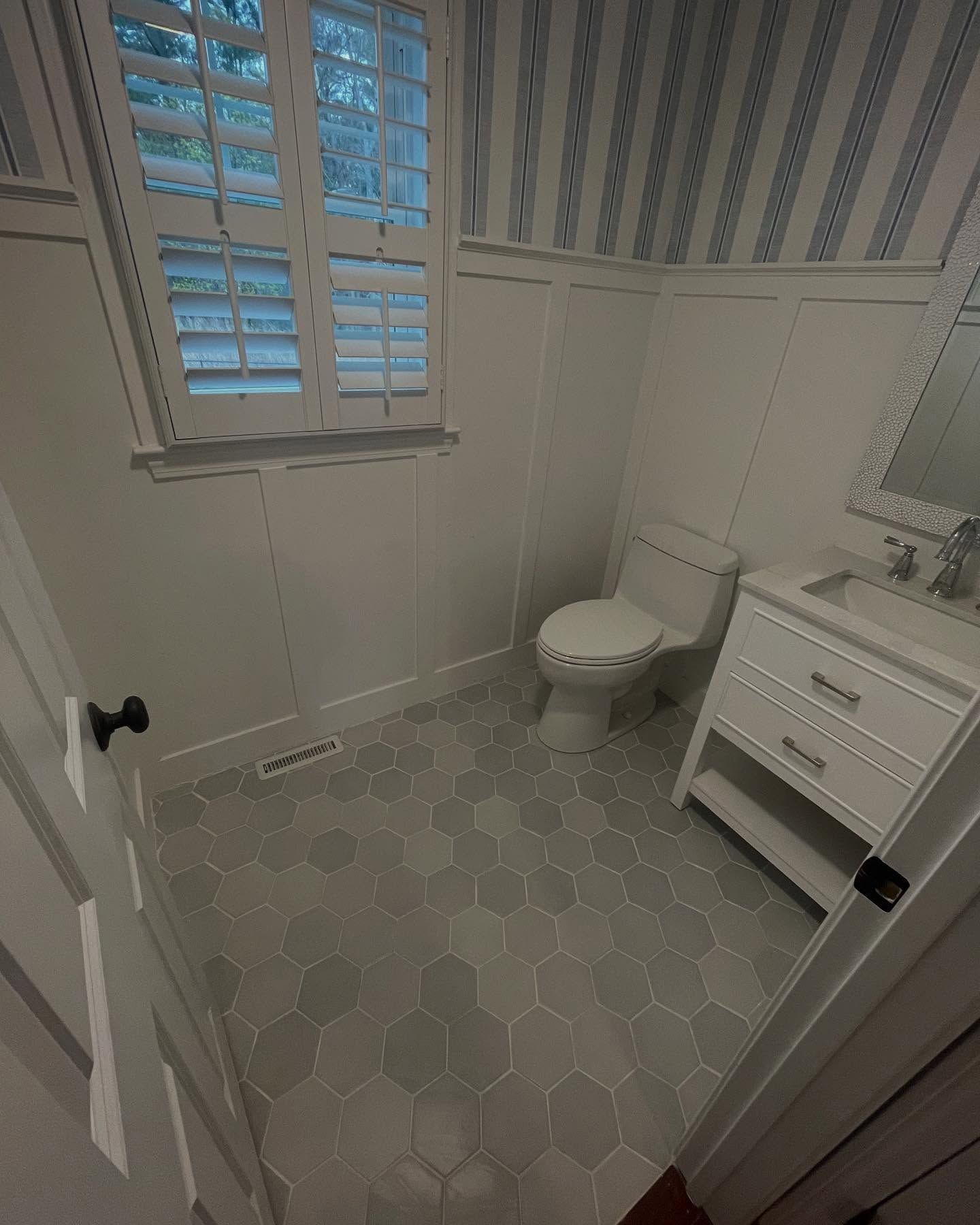 A small bathroom featuring white paneled walls, blue-and-white striped wallpaper, a white vanity, and gray hexagon flooring.