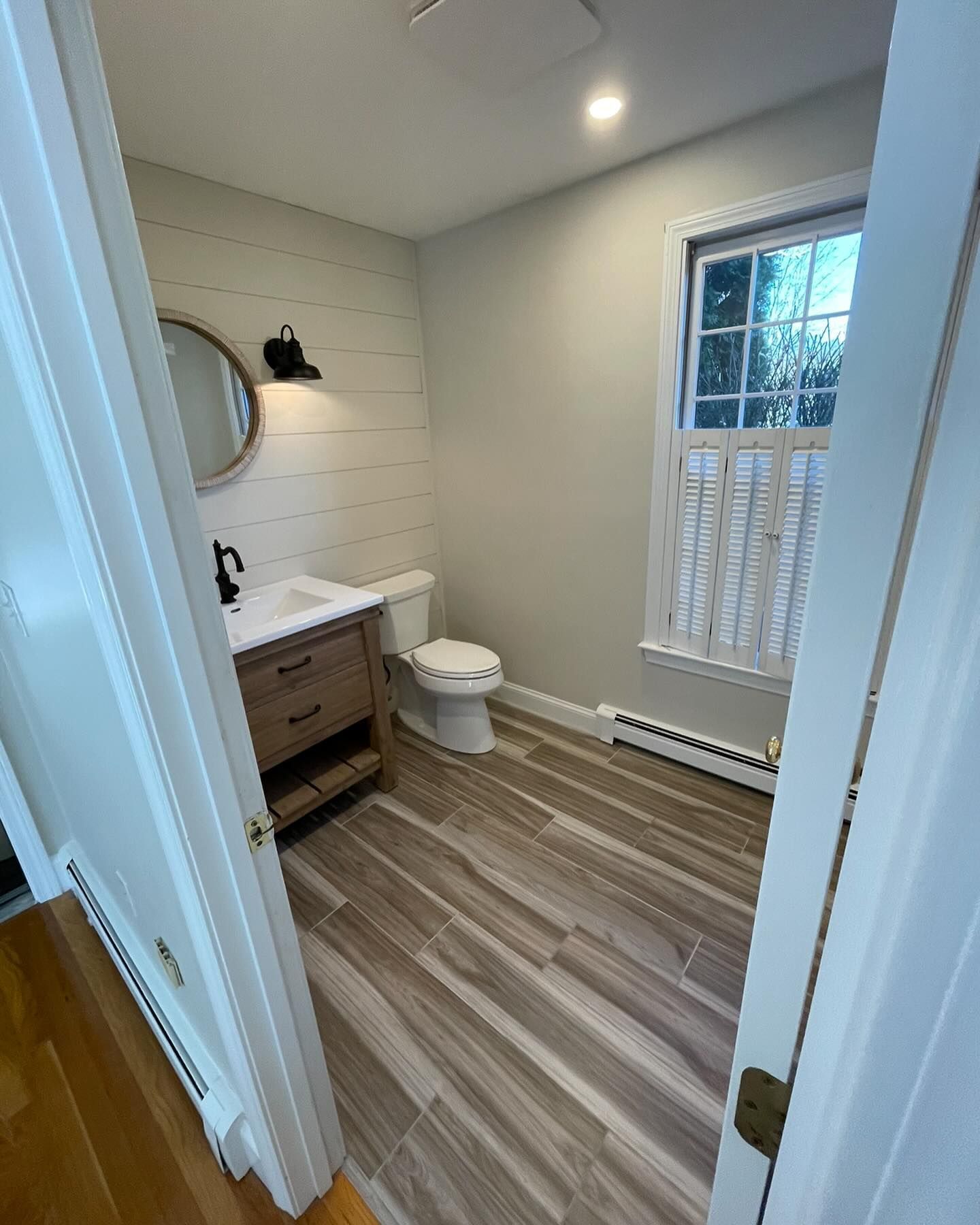 A small, bright bathroom with white shiplap walls, a wooden vanity, a round mirror, and wood-look tile flooring.