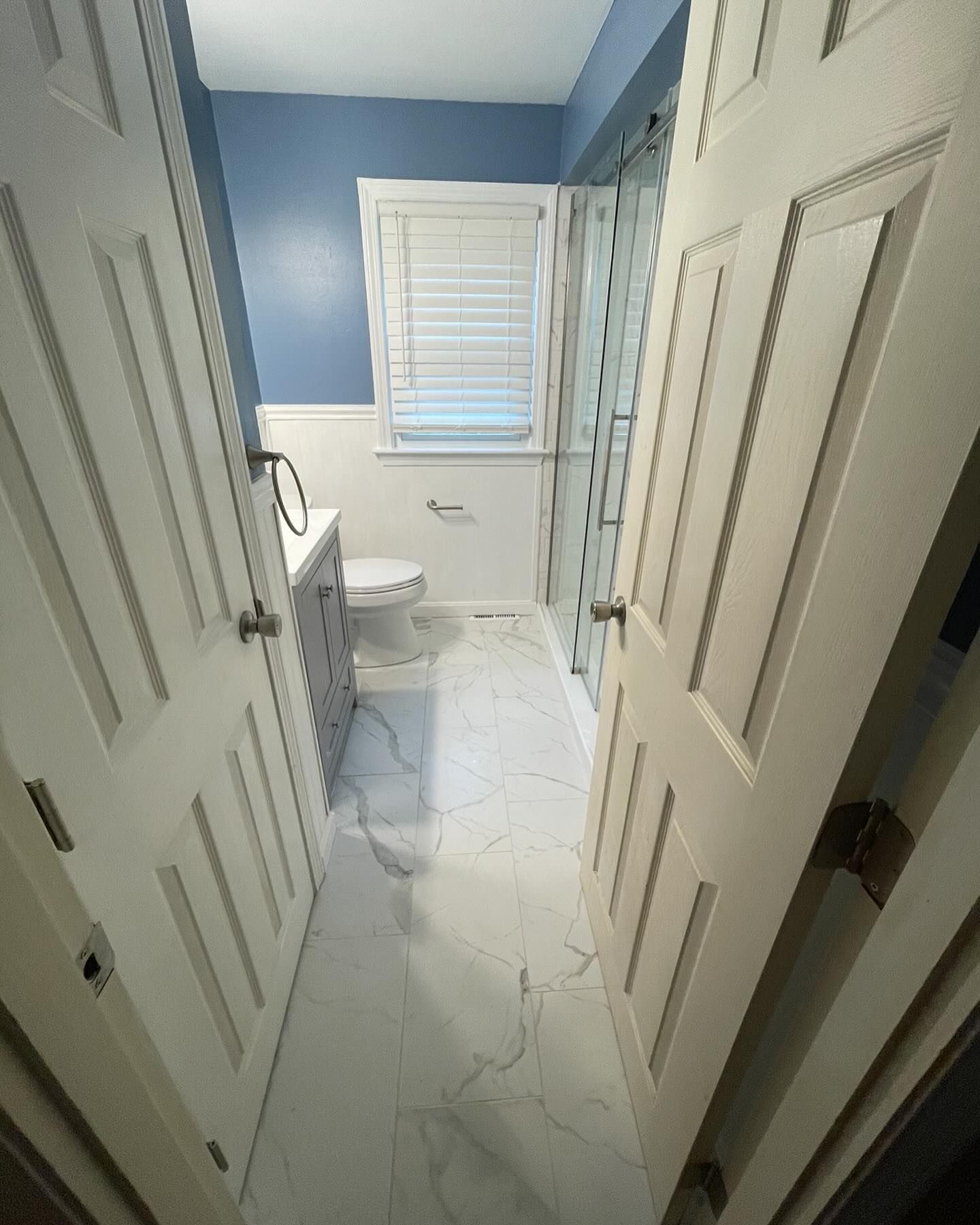 A view through a doorway into a small bathroom with white marble-patterned flooring, a white toilet, and blue walls.