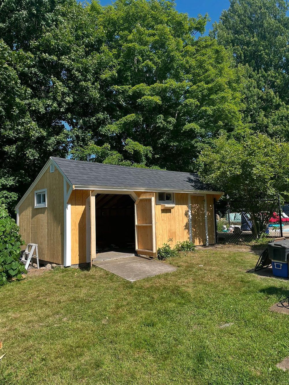 A light-colored wooden shed with a dark shingled roof stands on a grassy lawn in front of large, green trees.