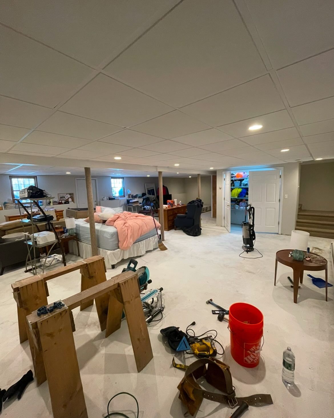 A basement under renovation with a bed, furniture, tools, and a red bucket on a light-colored carpeted floor.