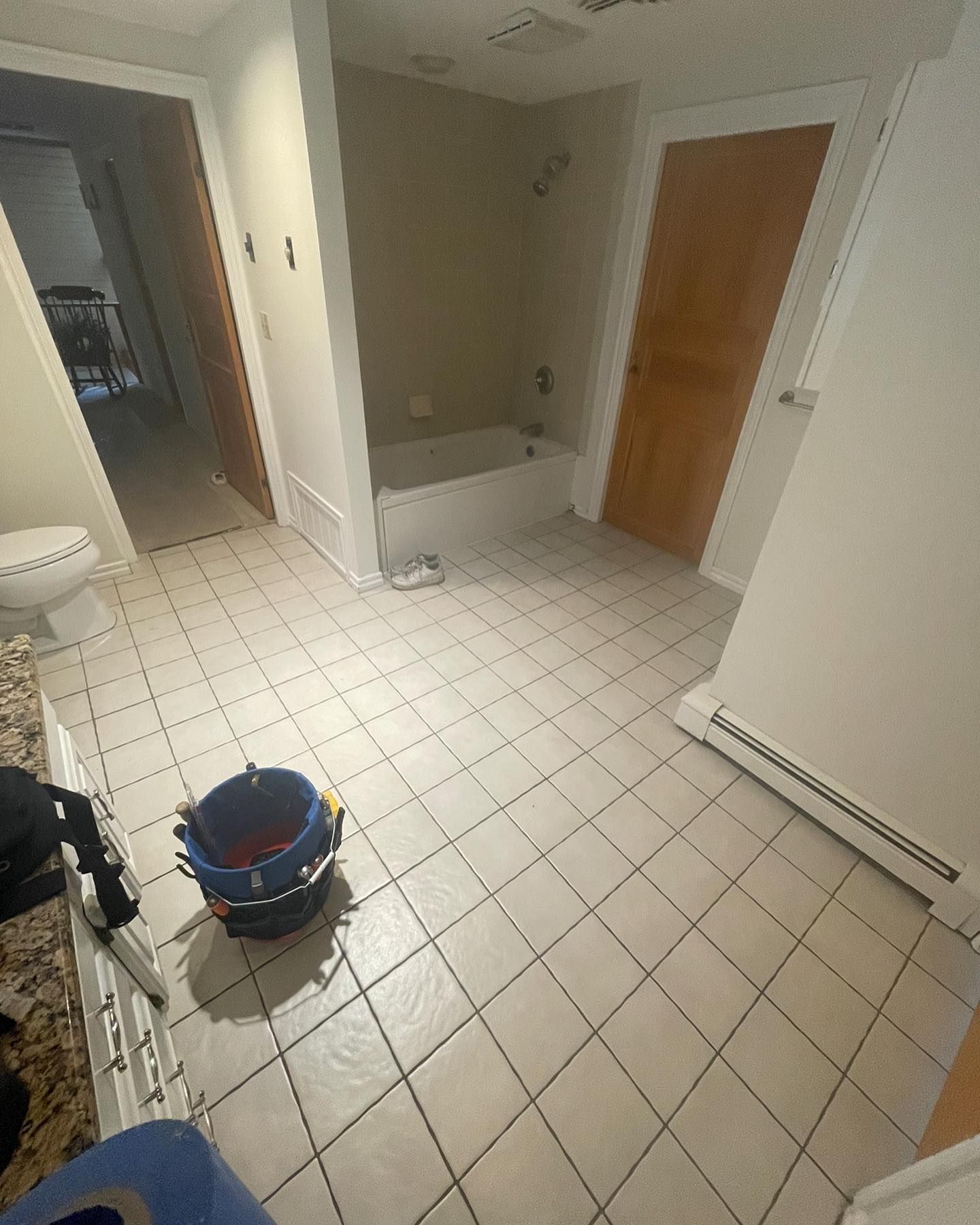 A bathroom with beige tile floors, a white bathtub, a wooden door, and a cleaning bucket on the floor.