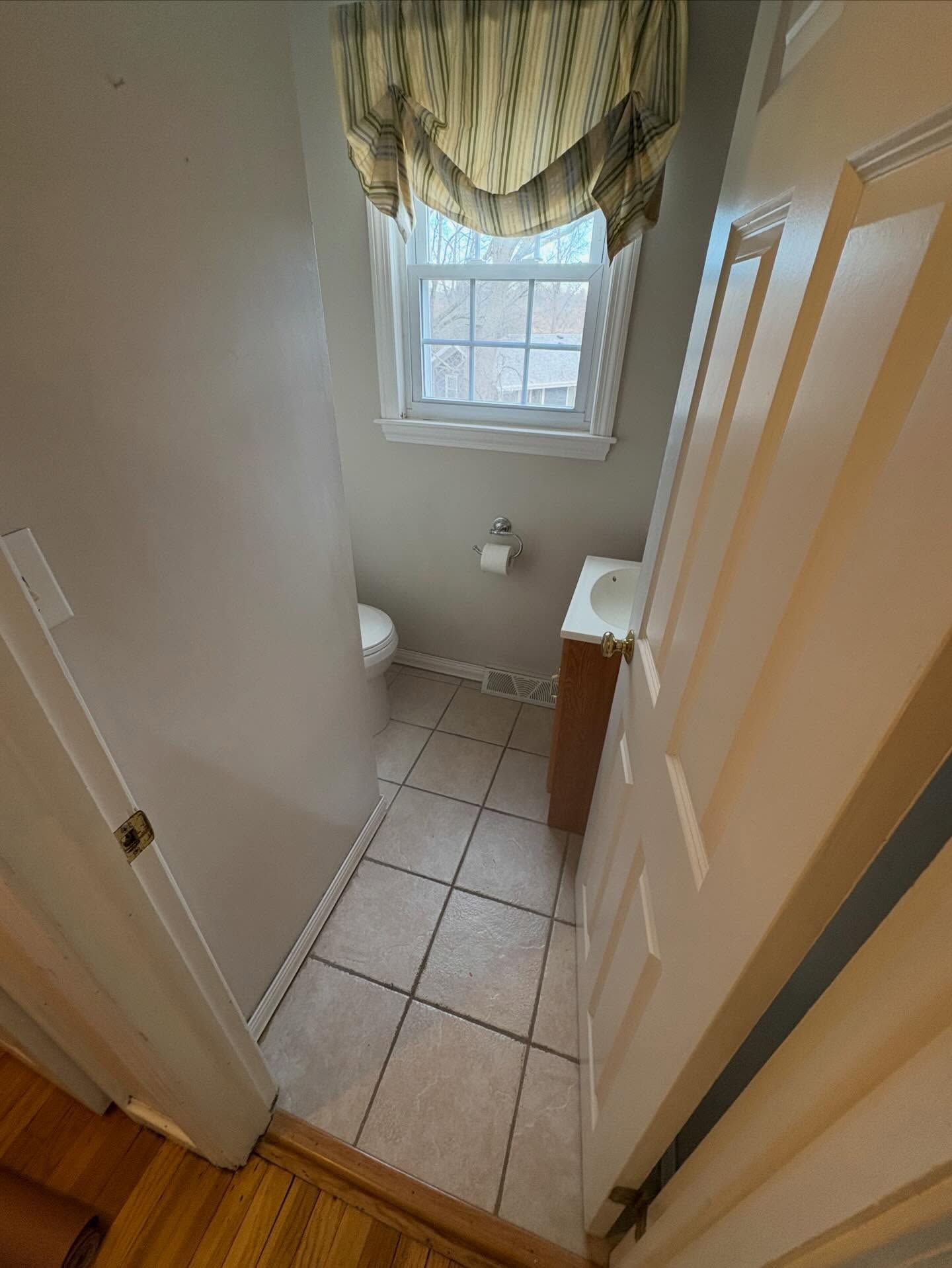 A narrow powder room with a tiled floor, small vanity, toilet, and a window with a valance, viewed through an open doorway.