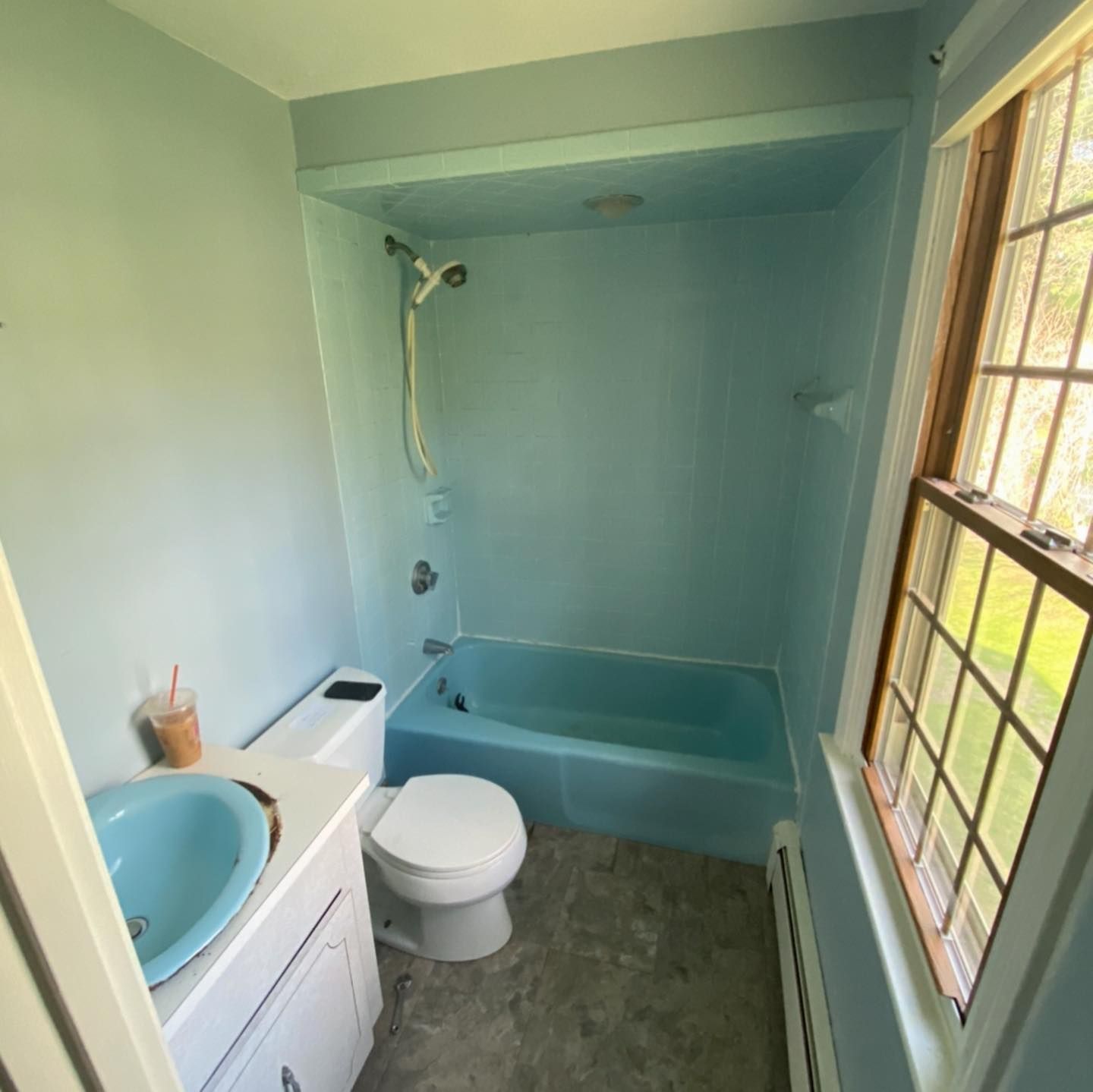 A bathroom with light blue walls, a matching blue sink and tub, a white toilet, and a window with wooden trim.