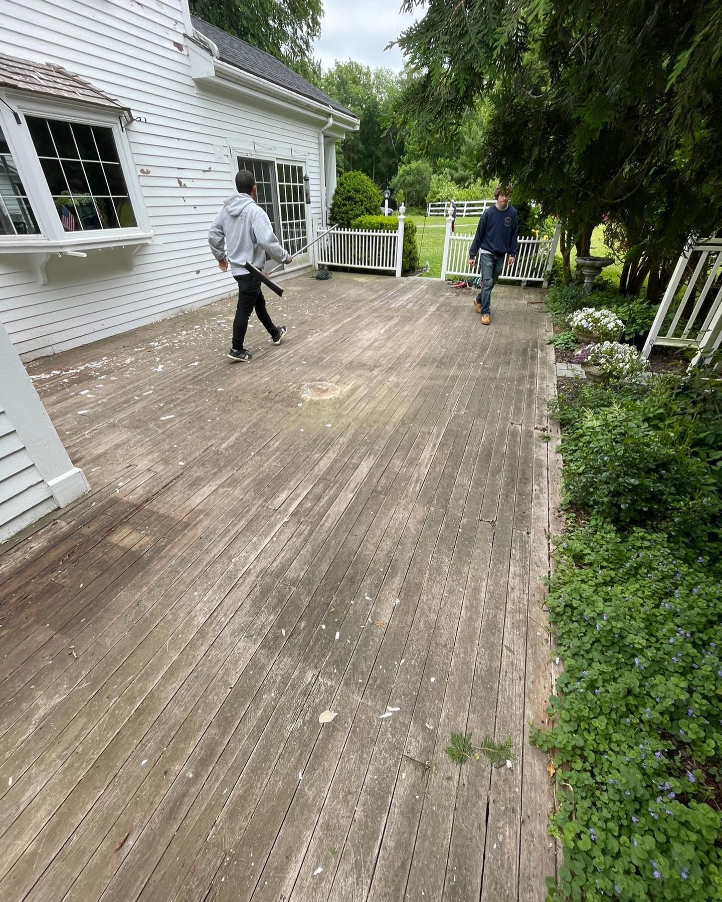 Two people walk across a weathered wooden deck attached to a white house with white railing and lush greenery nearby.