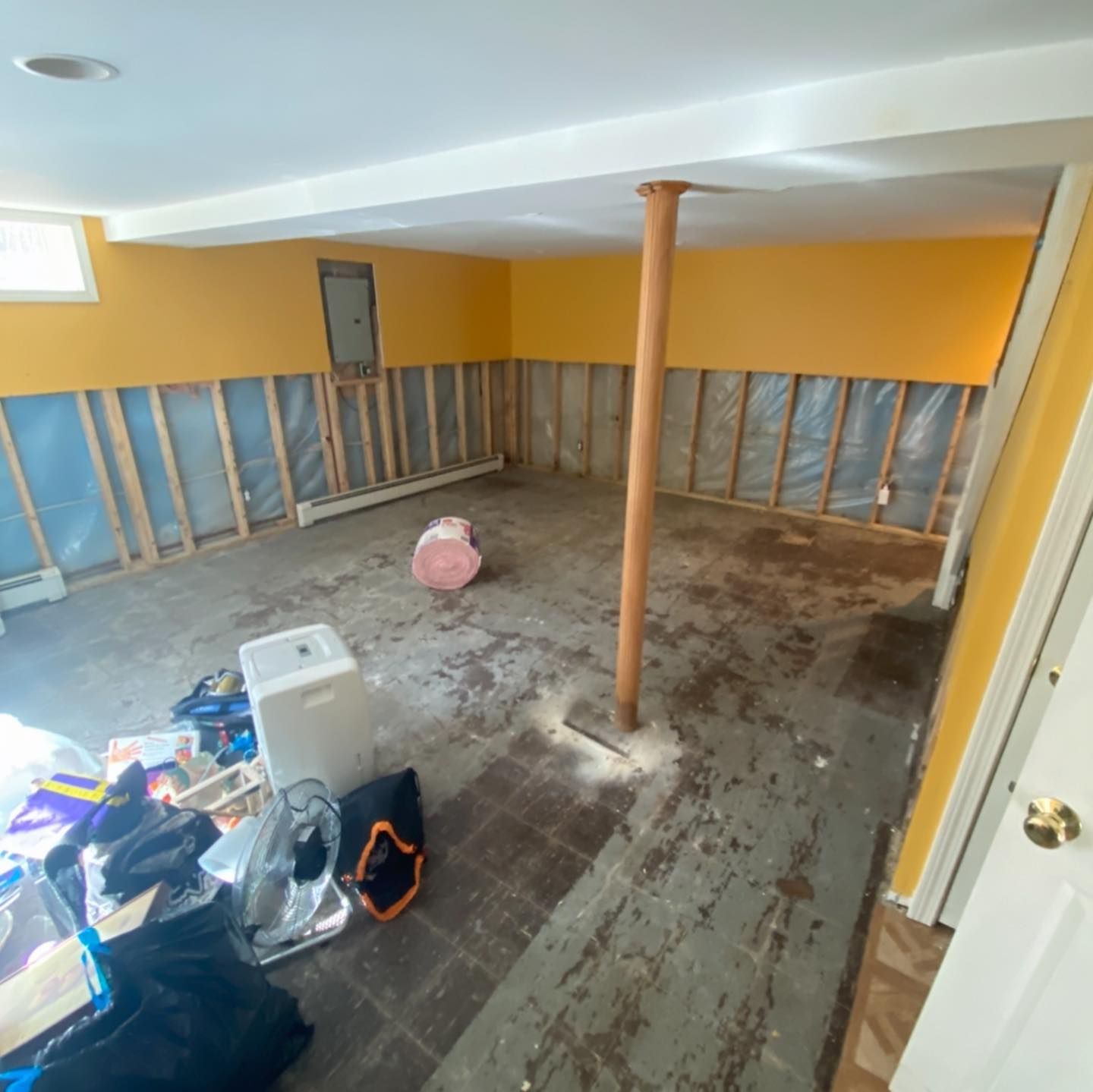 Basement room undergoing renovation with yellow painted walls, exposed wooden studs, a metal support pole, and messy floor.