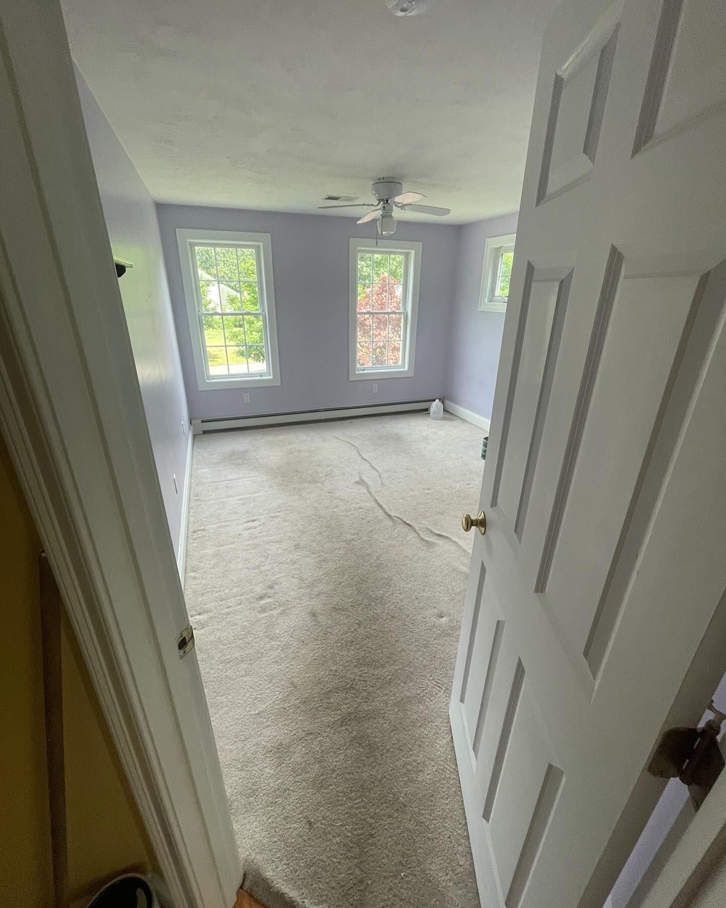 An open white door reveals a sparsely furnished, light purple bedroom with beige wall-to-wall carpeting and two windows.