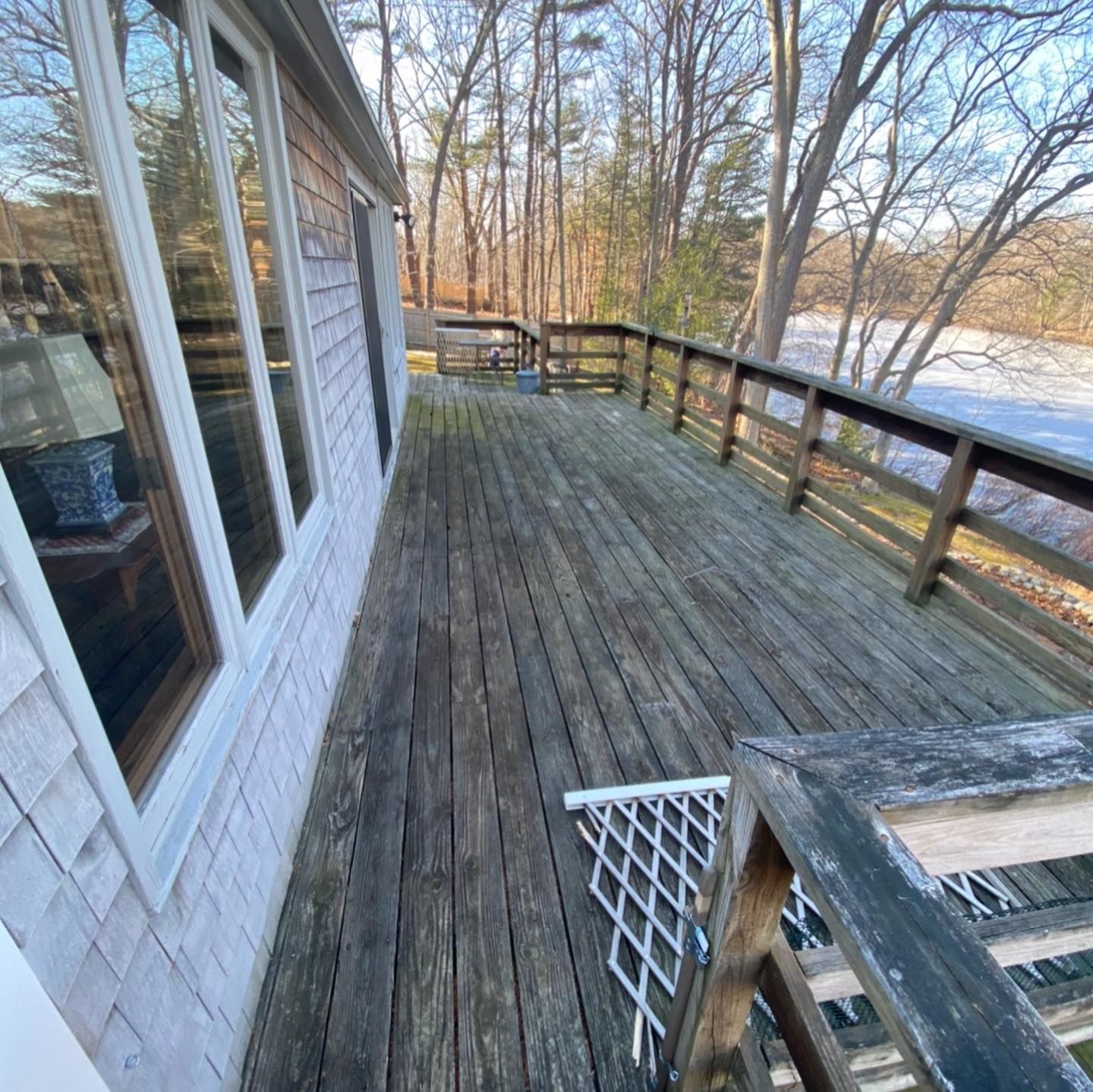 A weathered wooden deck runs alongside a shingled house, overlooking trees and a frozen body of water.