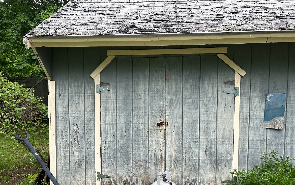 A weathered gray wooden shed with a faded shingled roof and cream-colored trim, set against a backdrop of green trees.