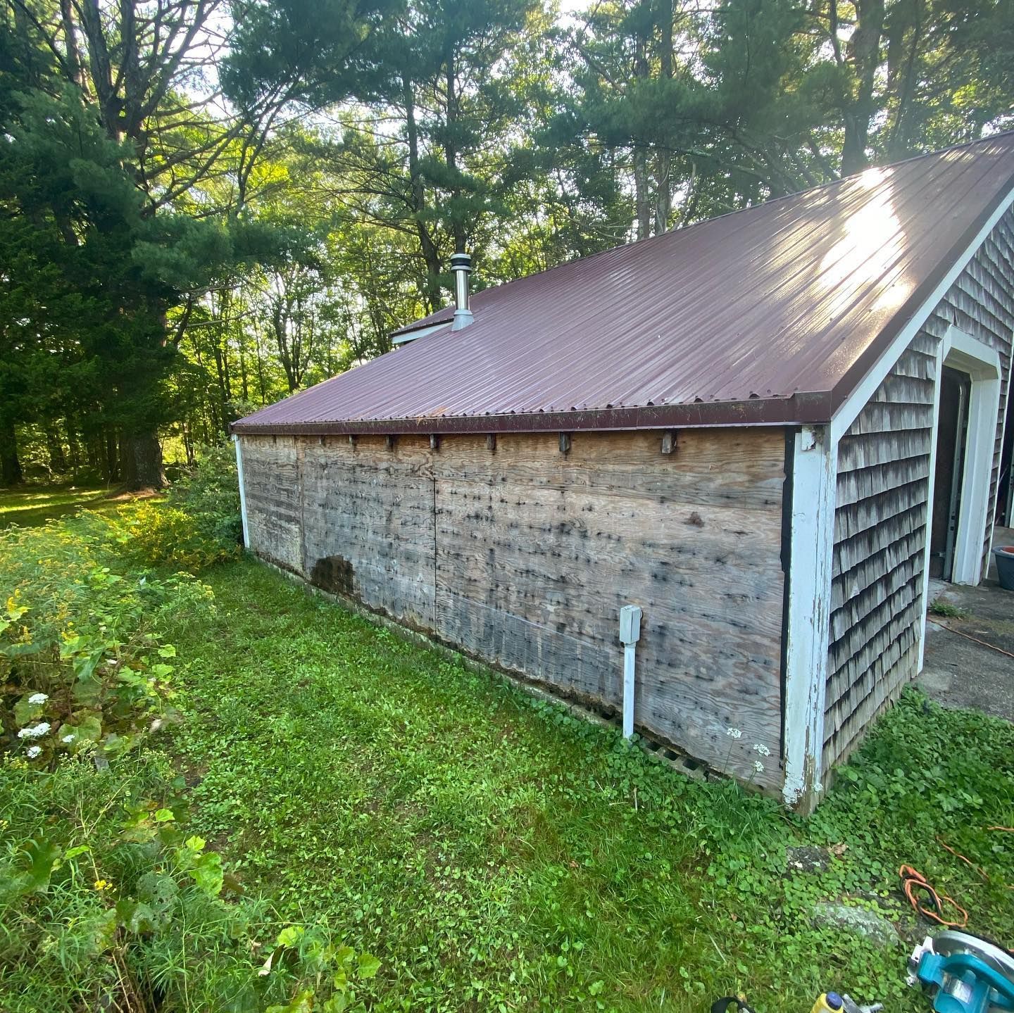 A side view of a shed with a reddish-brown metal roof and weathered wooden walls, set against trees and a grassy lawn.