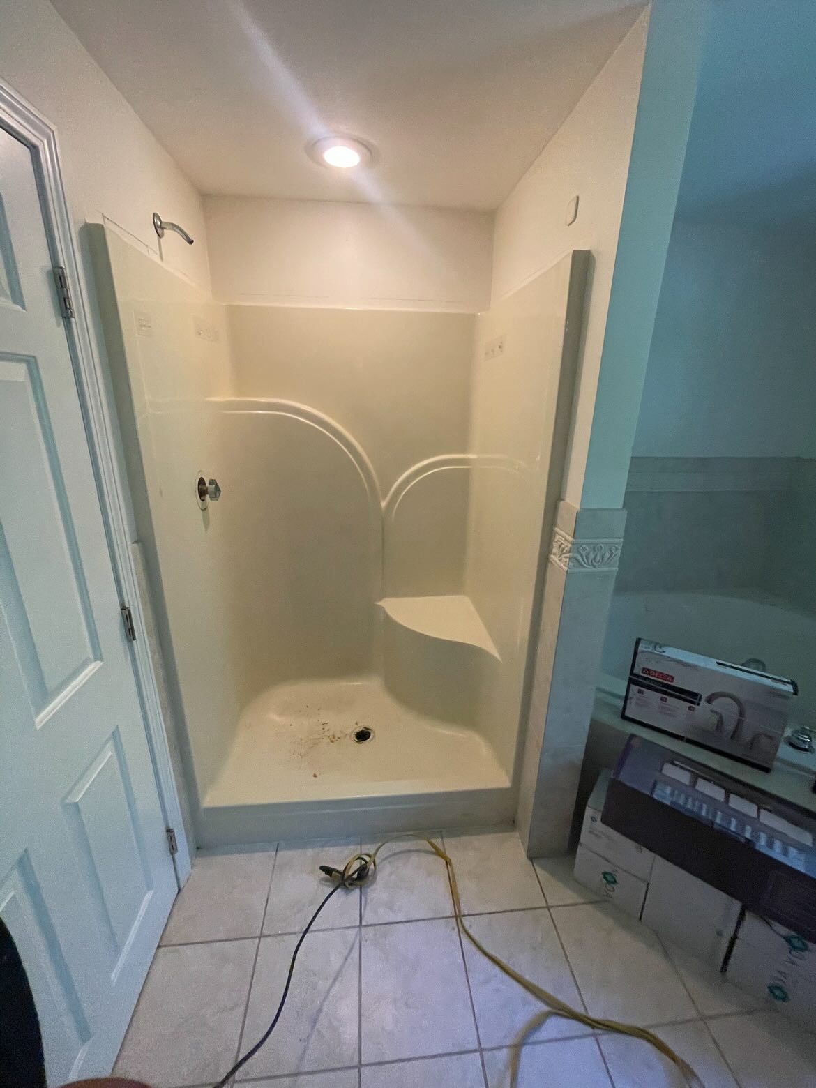 A beige pre-fabricated shower stall with a built-in seat, set in a tiled bathroom with a doorway and nearby vanity.