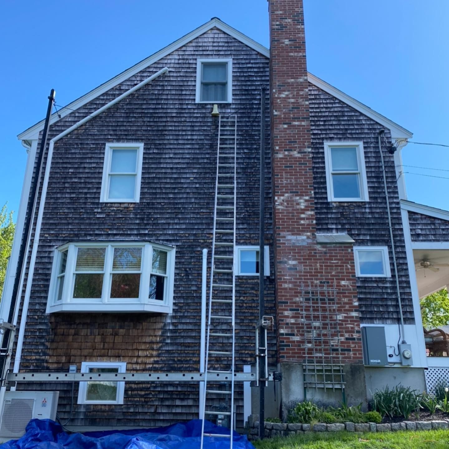 A two-story house with weathered wood shingles, a brick chimney, multiple windows, and a tall ladder leaning against it.
