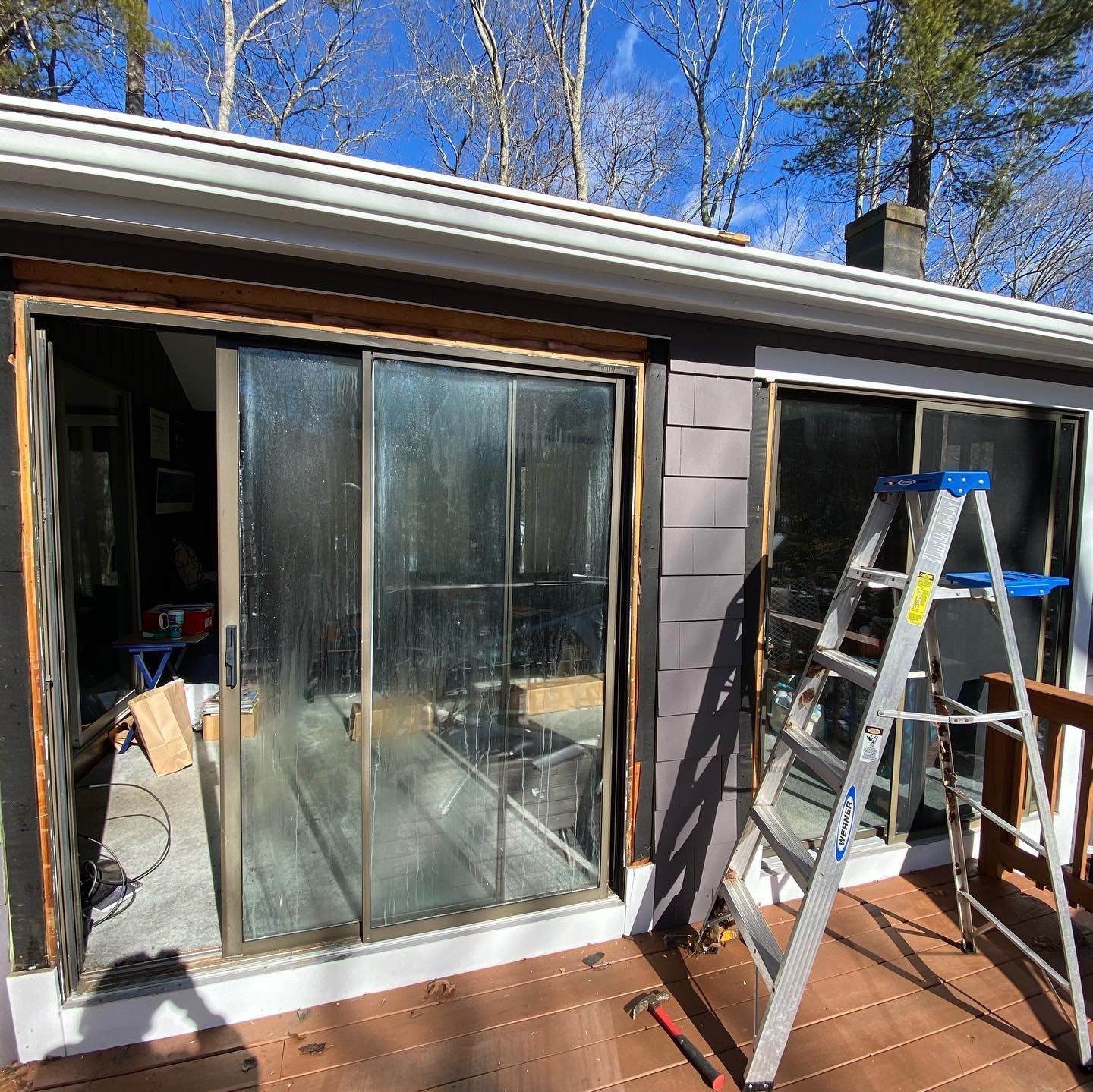 A sliding glass door under renovation on a deck with a silver ladder leaning against the exterior siding.