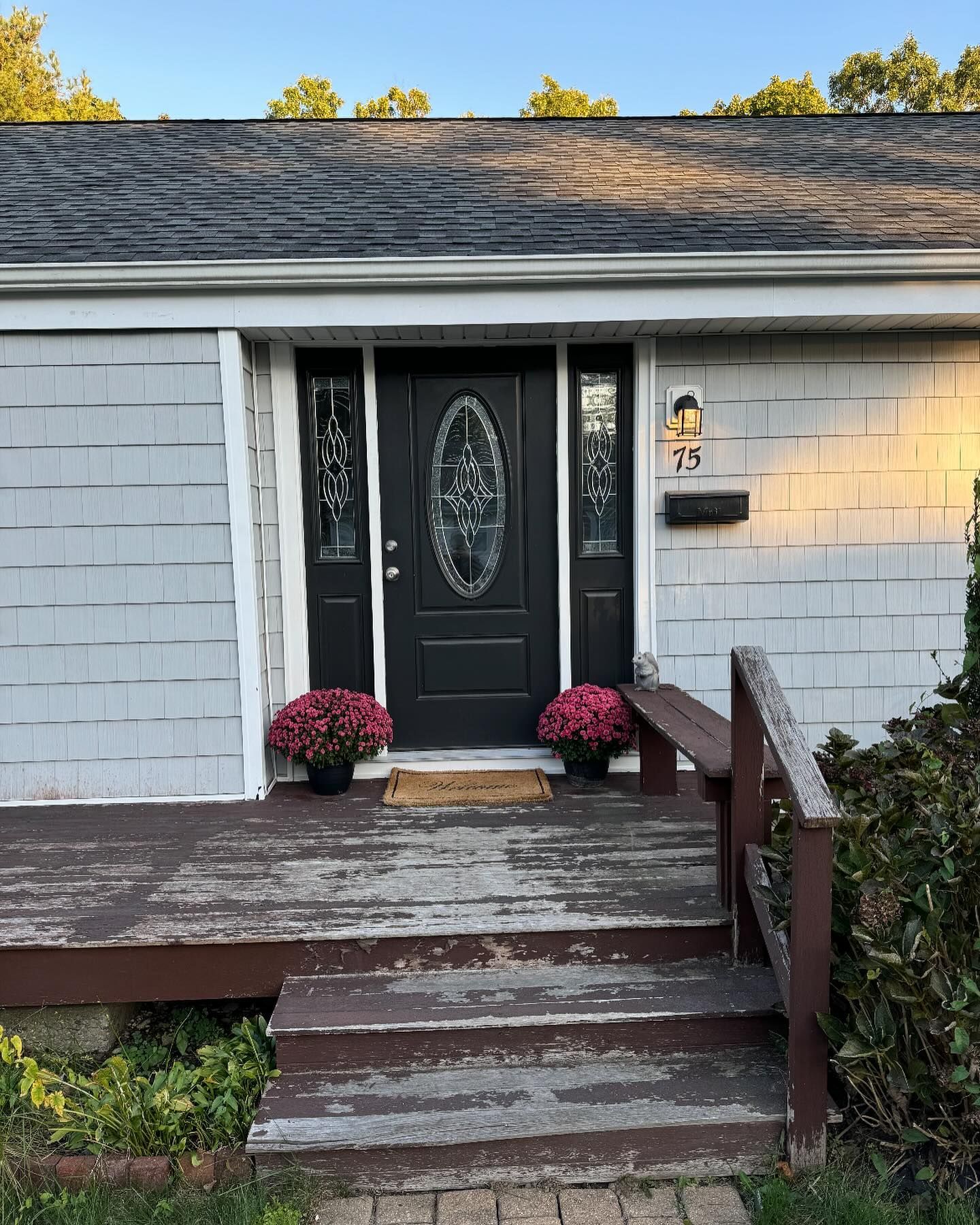 A front porch with weathered dark brown steps leads to a black front door flanked by sidelights and pink potted mums.