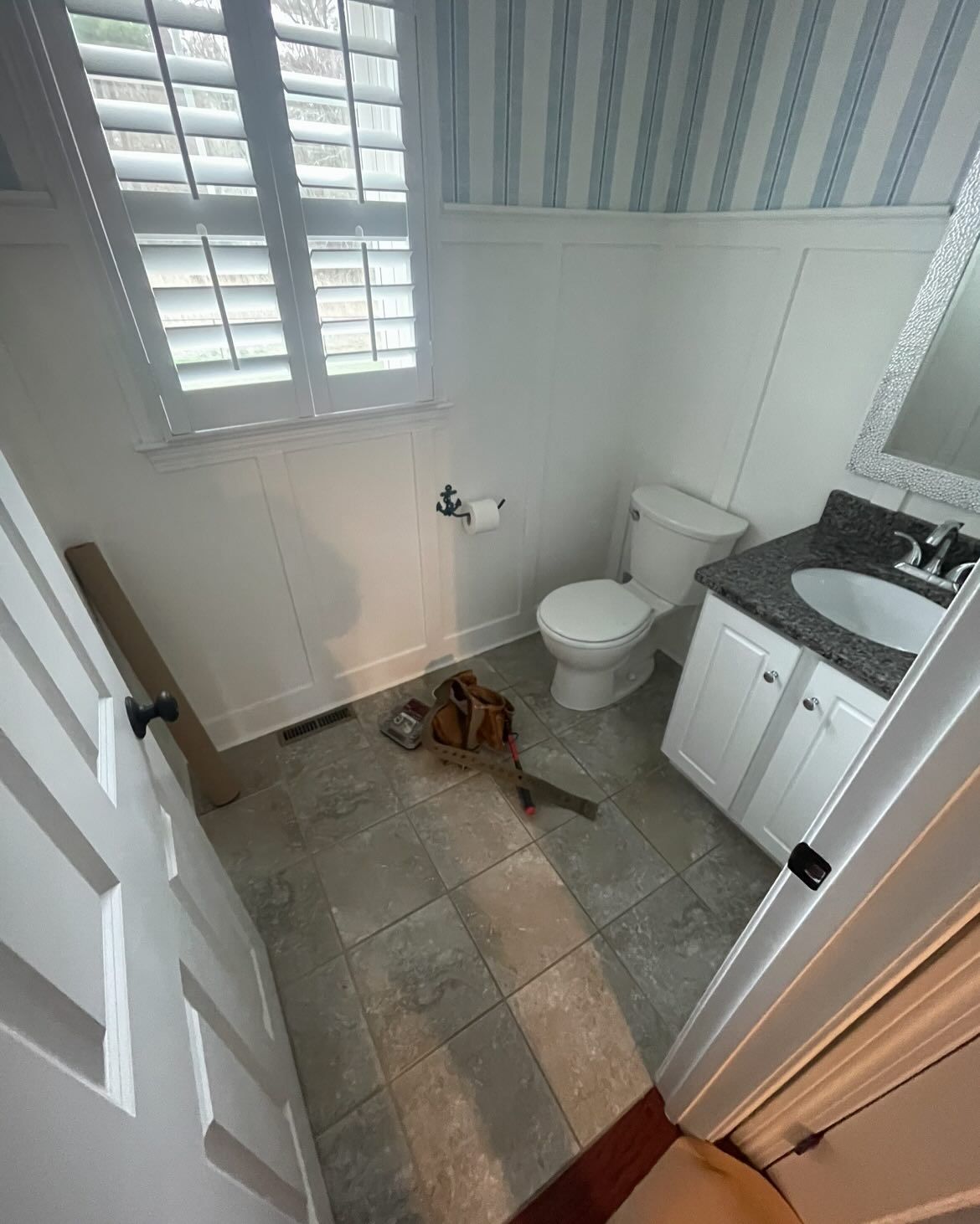 A small bathroom with a toilet, a vanity with a dark stone counter, tile flooring, and blue-and-white wallpaper.