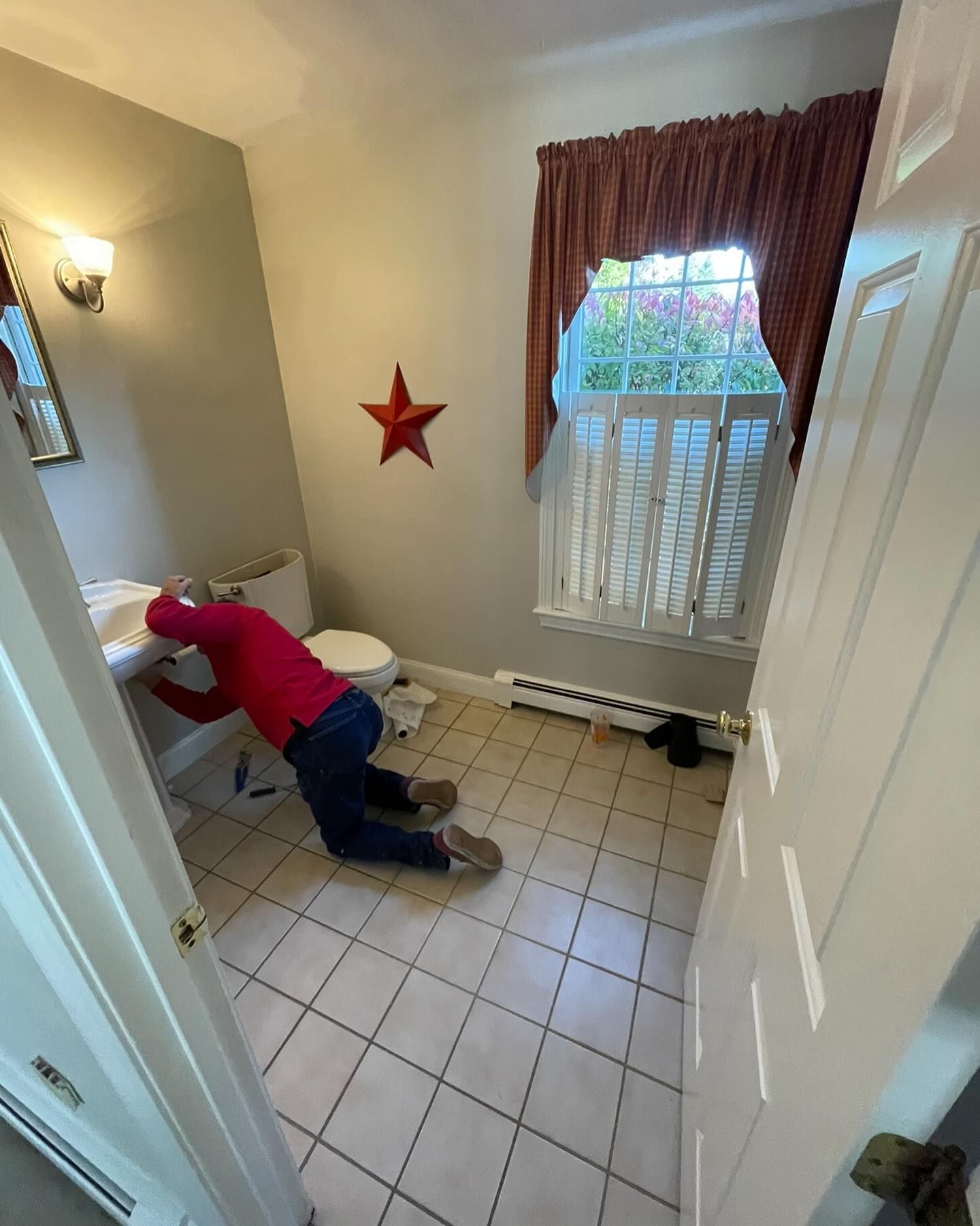 A person in a red shirt kneels on a tiled bathroom floor while working on plumbing under the sink.