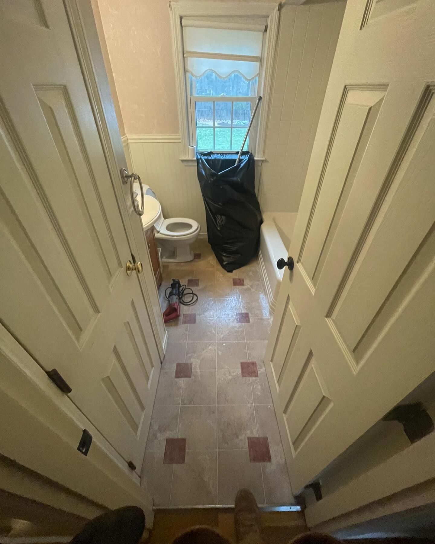 A view from a doorway into a small bathroom with light tile flooring, a toilet, a window, and a large black trash bag.