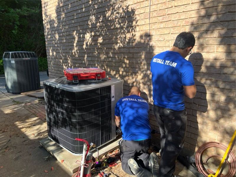 Two men in blue shirts repair an AC unit next to a brick wall, with tools and copper tubing visible.