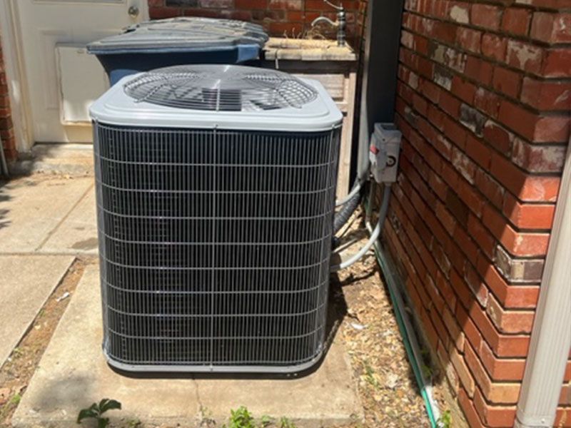 An air conditioning unit outside, next to a brick wall and a trash can.