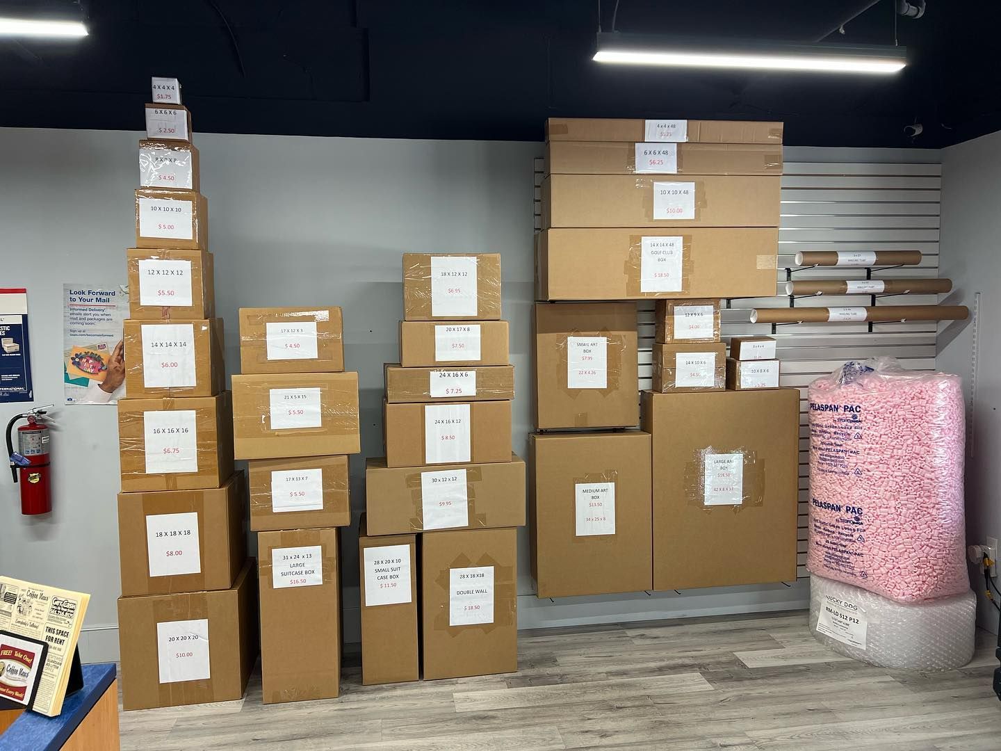 A stack of various cardboard shipping boxes of different sizes and a large bag of pink packing peanuts inside a store.