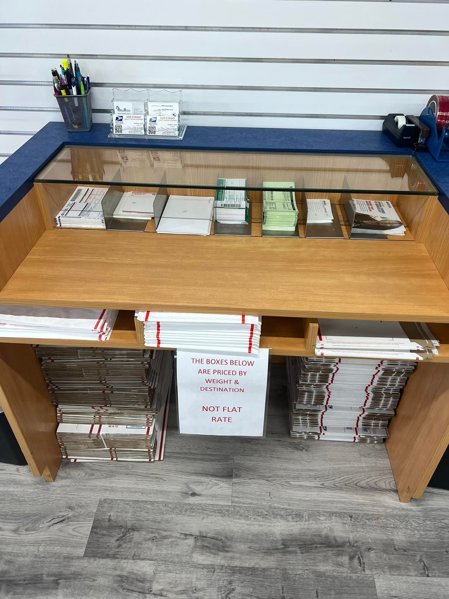 A wooden service counter with glass-topped bins filled with paper forms and stacks of white mailers underneath.