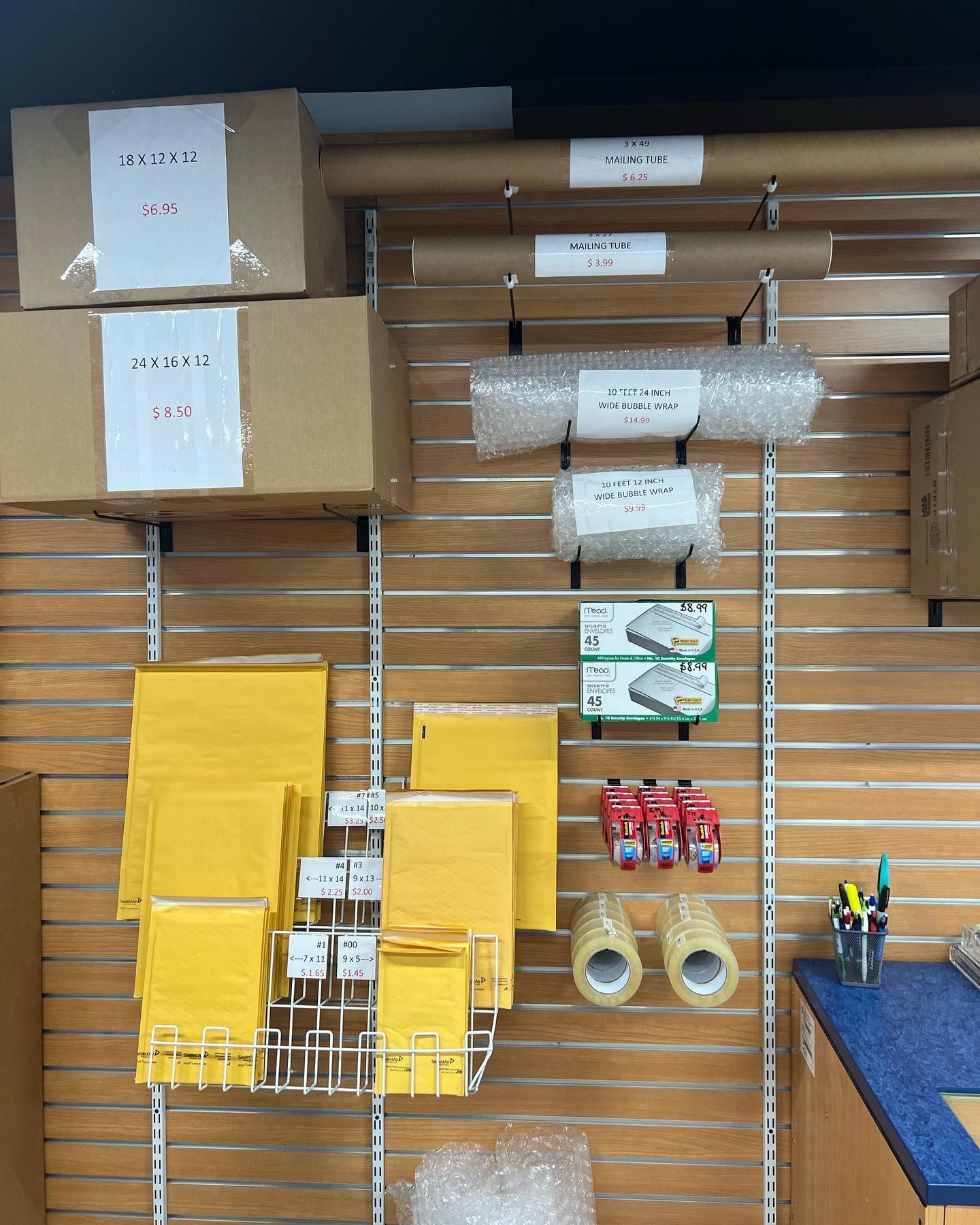 Retail display wall with cardboard shipping boxes, mailing envelopes, packing paper, bubble wrap, and adhesive tape rolls.