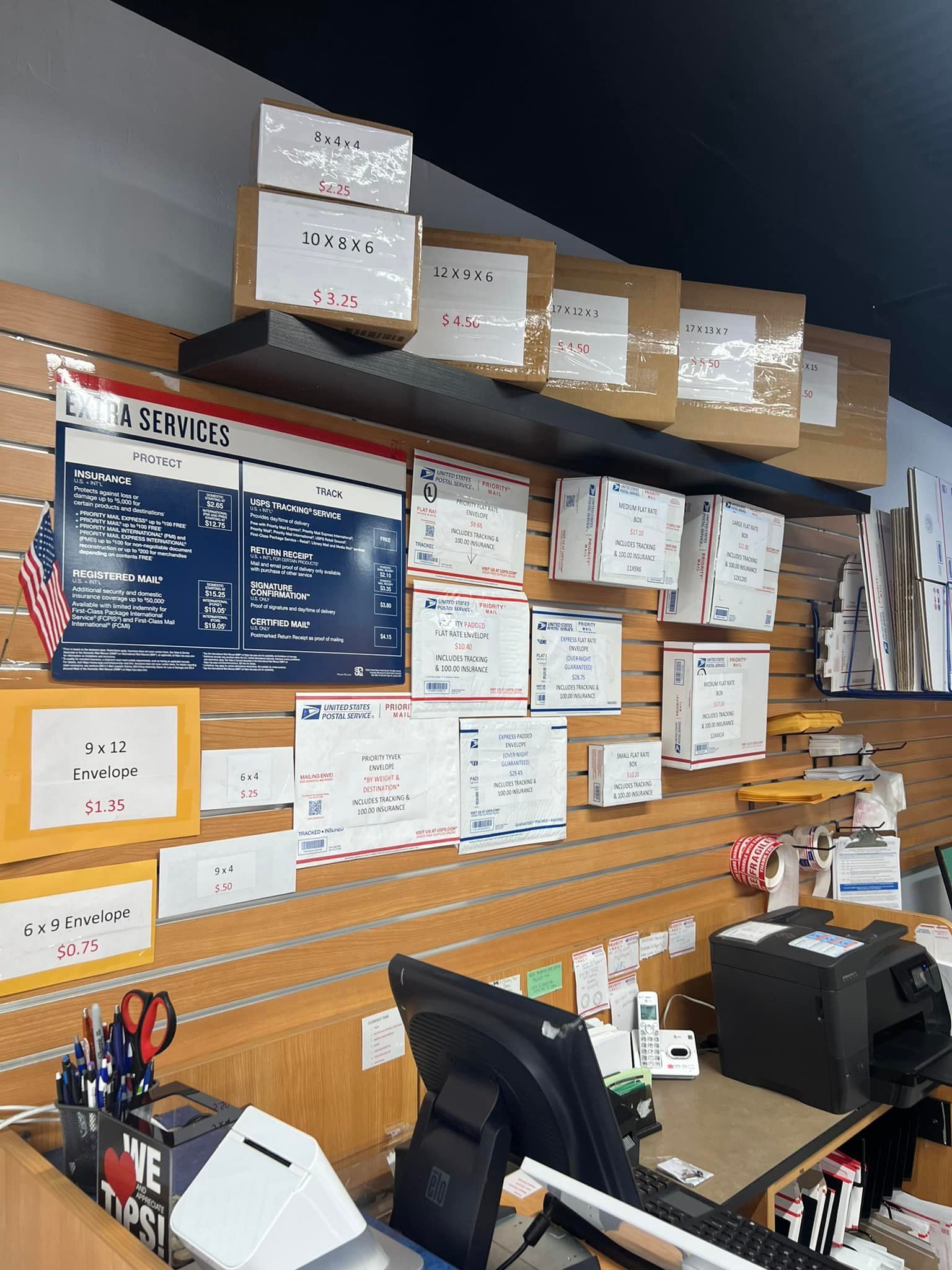 A service counter at a United States Post Office, featuring shipping boxes, price charts, and forms on a slat wall.