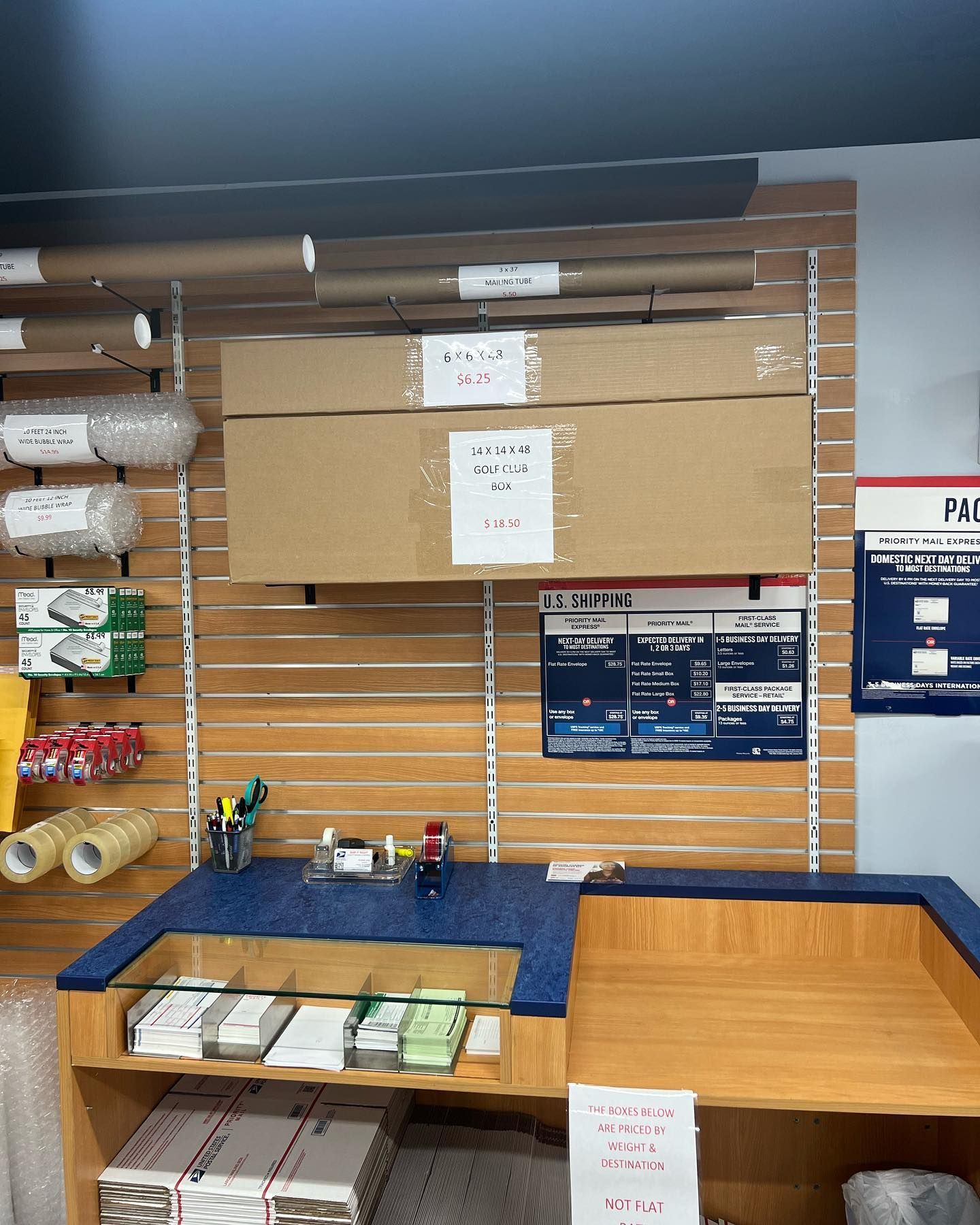 A service counter at a shipping or mailing store with packing supplies, cardboard boxes, and forms on a slat wall.