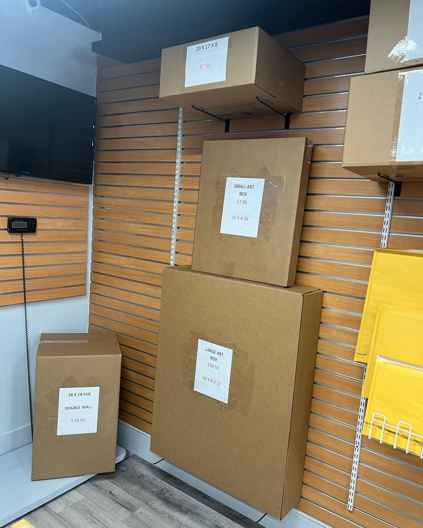 Cardboard boxes of various sizes with shipping labels attached, displayed on a wooden slat wall in a shipping store.