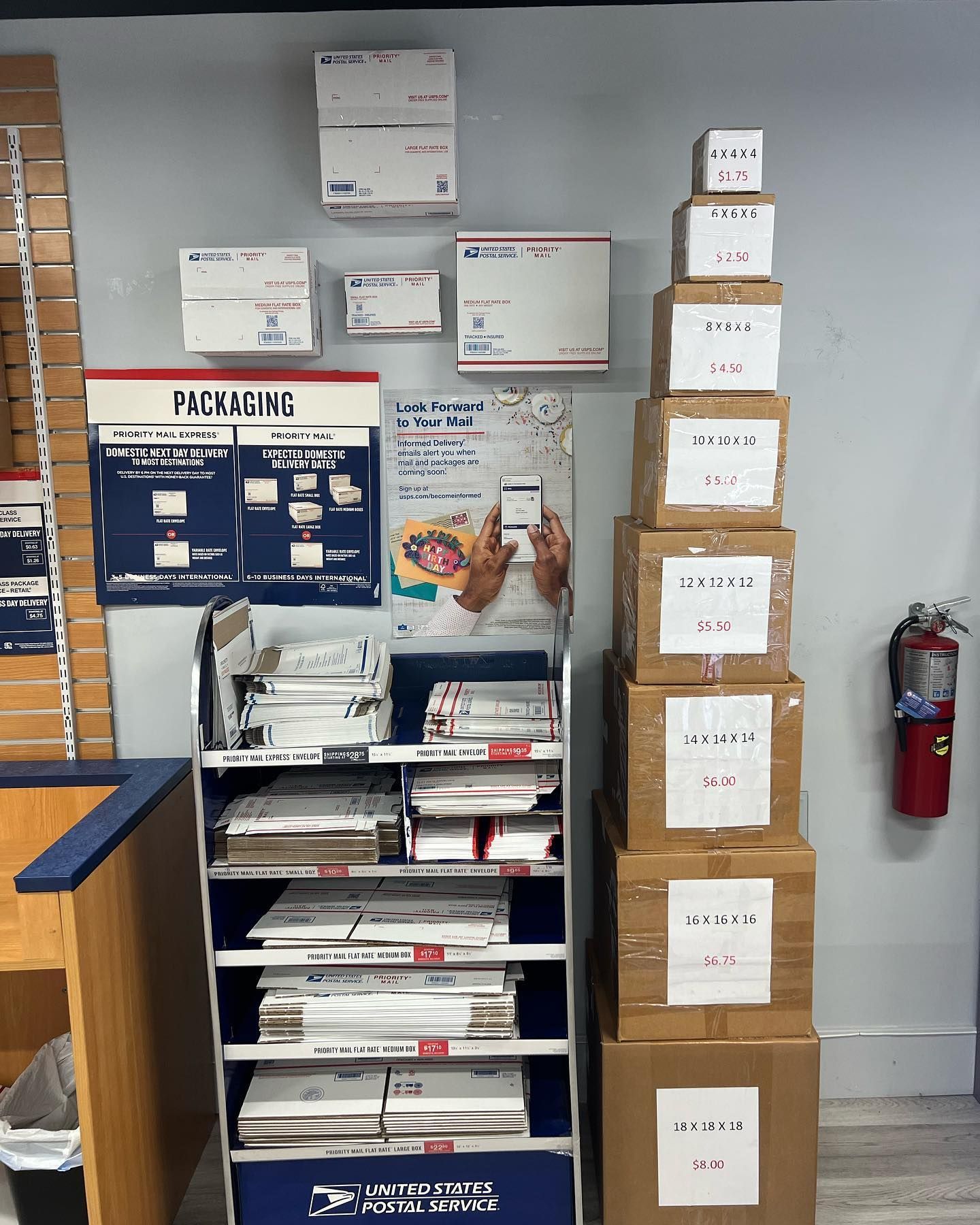 A post office wall with postal packaging supplies on a shelf and a tower of stacked shipping boxes of varying sizes.