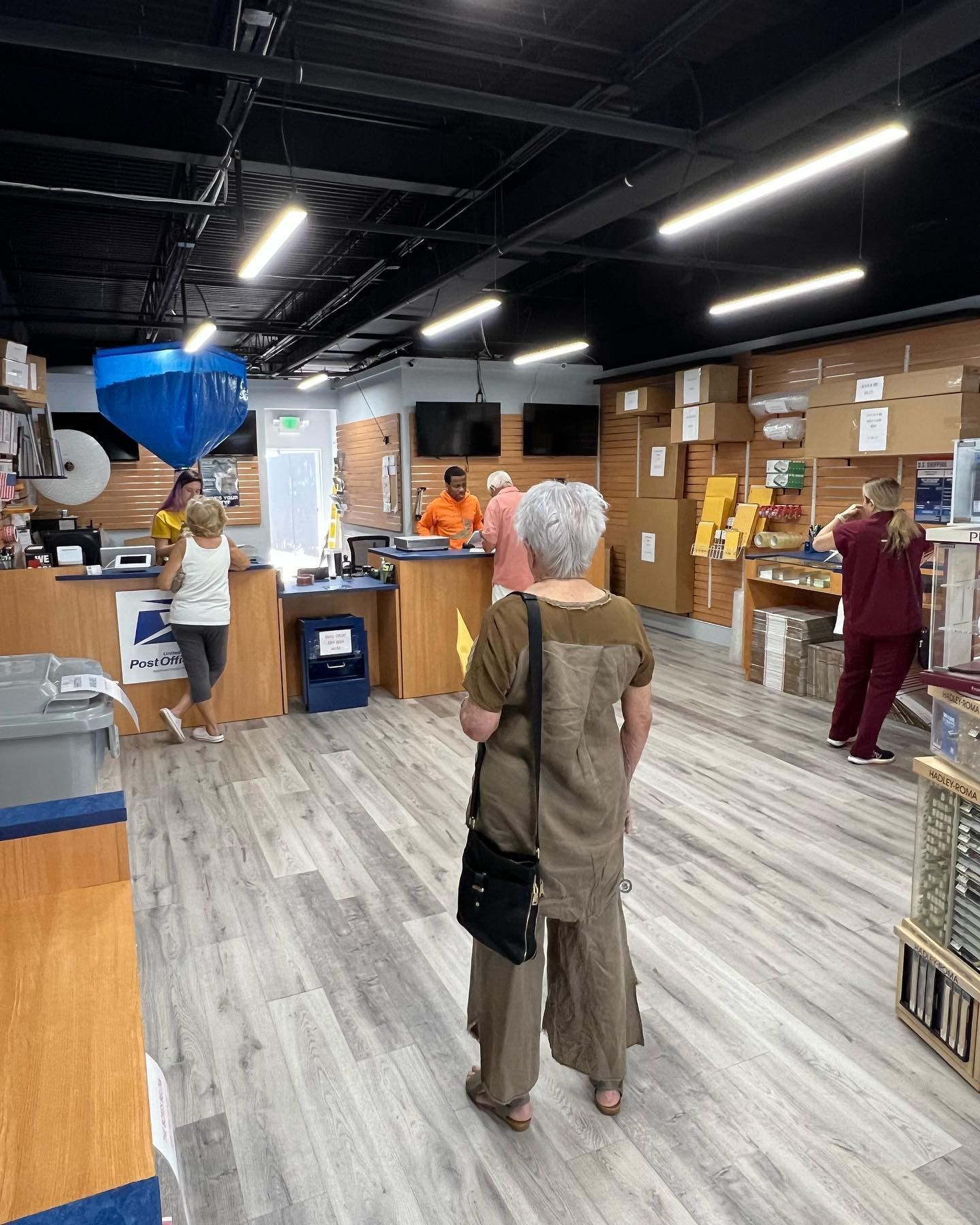 Patrons stand in line at a shipping store counter with wood-paneled walls and shelves stocked with boxes.