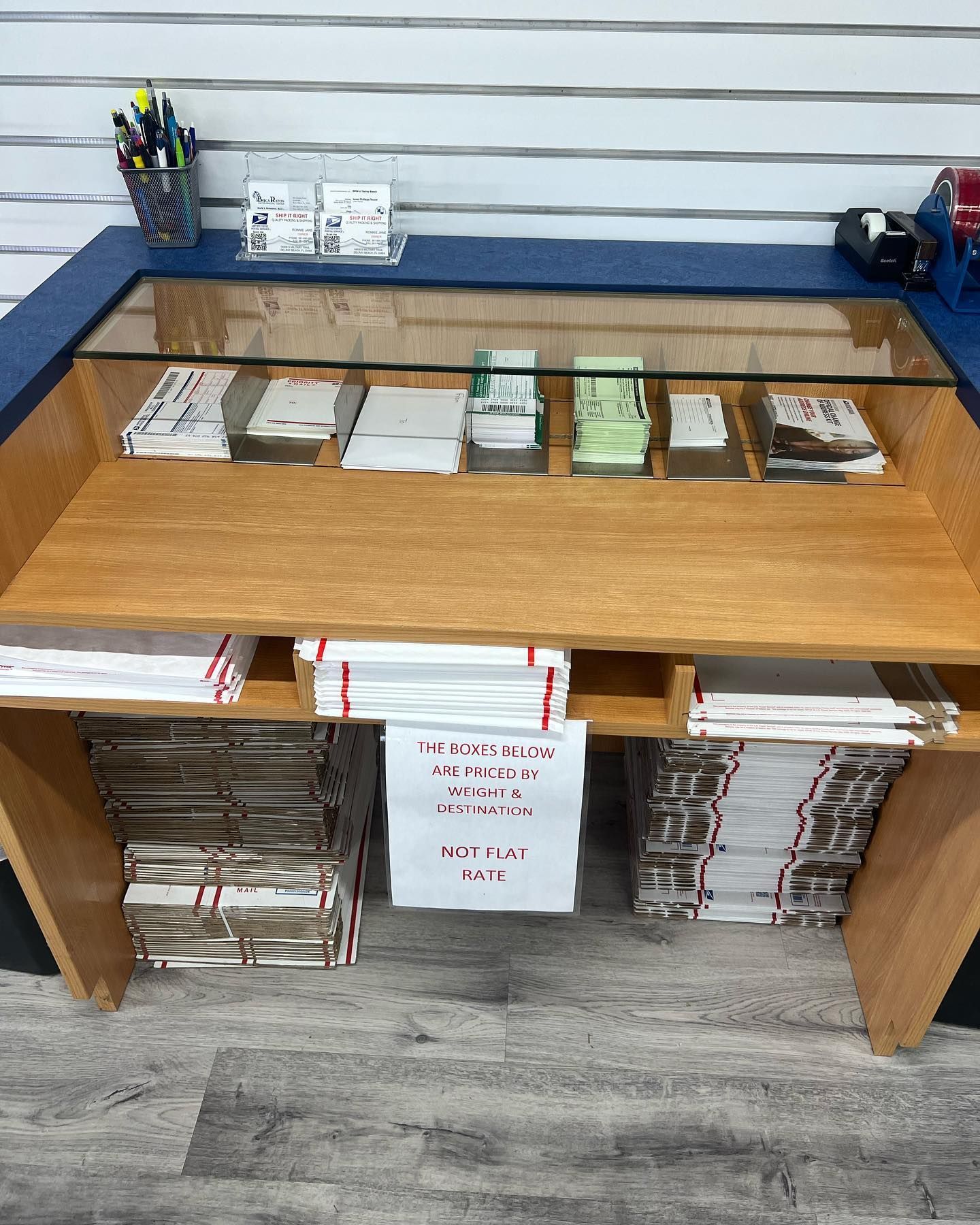 A wooden customer service counter with a glass top, stacked paperwork, and a sign asking customers to wait for an employee.