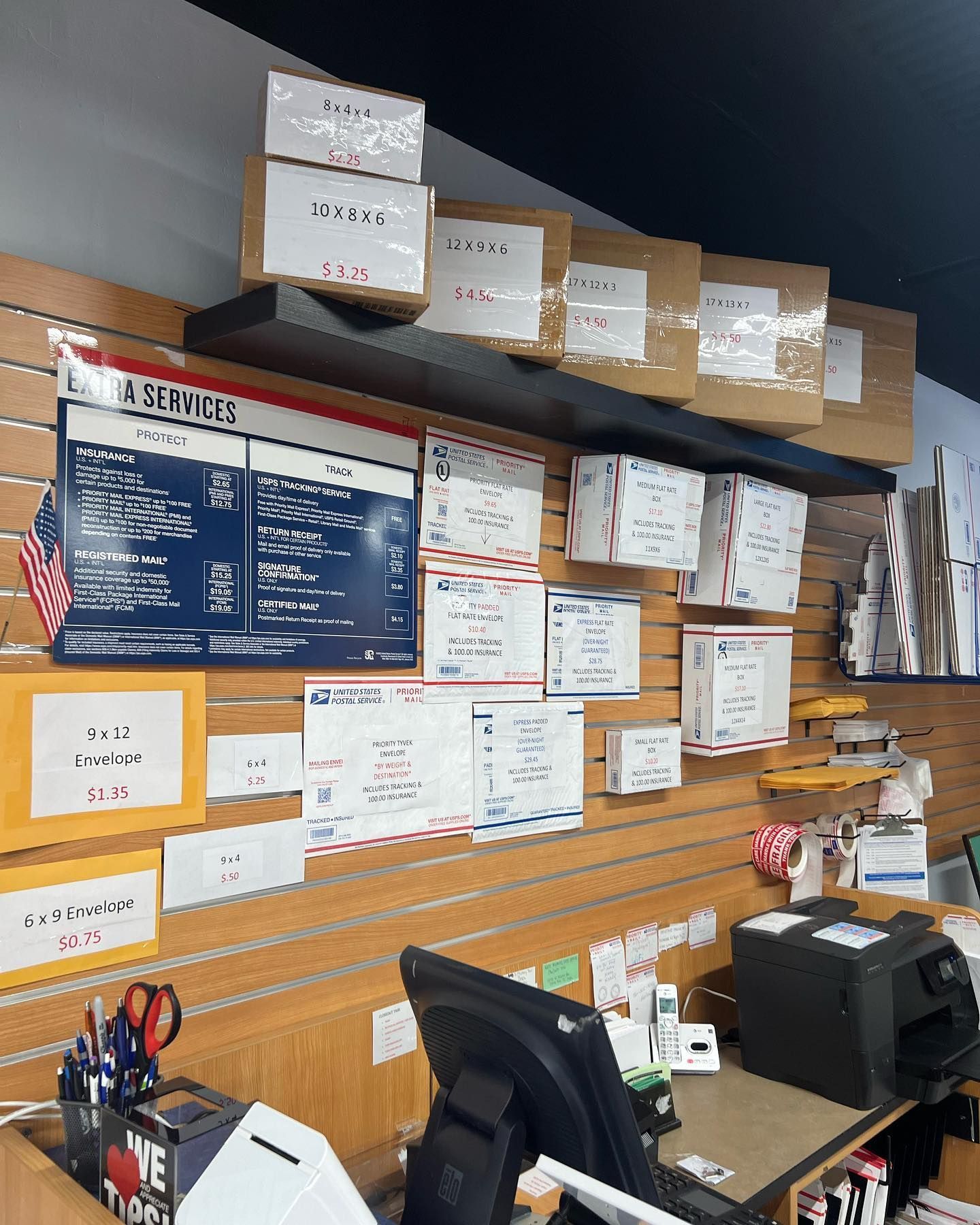 A retail shipping counter with shelves stocked with various cardboard boxes, a services sign, and a black computer monitor.