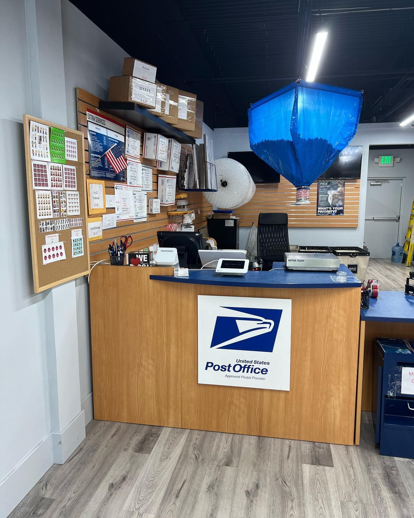 A wooden service counter at a United States Post Office with a blue hanging sign and a cork bulletin board on the left.