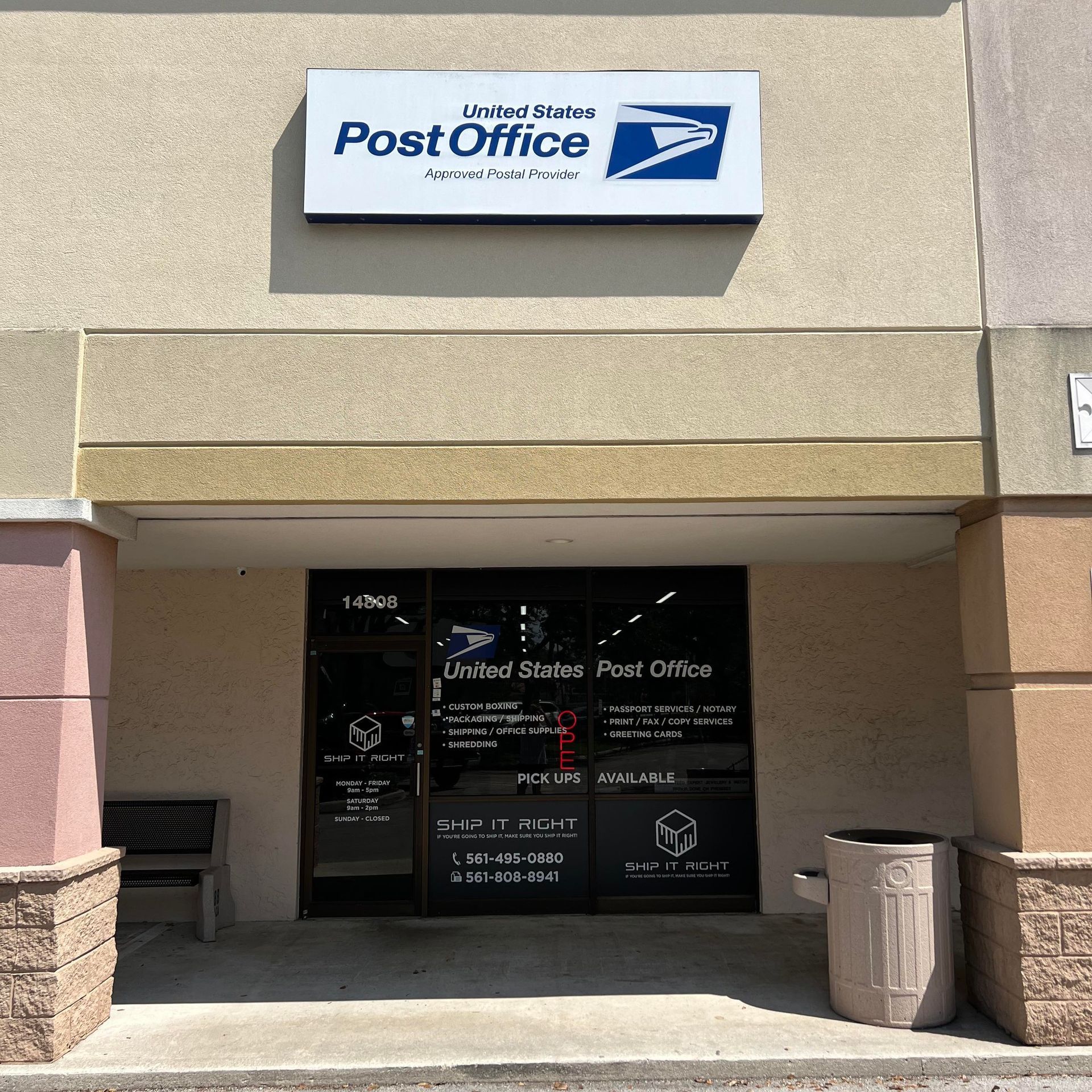 A United States Post Office storefront with a large white sign above the glass entrance doors and a trash bin outside.