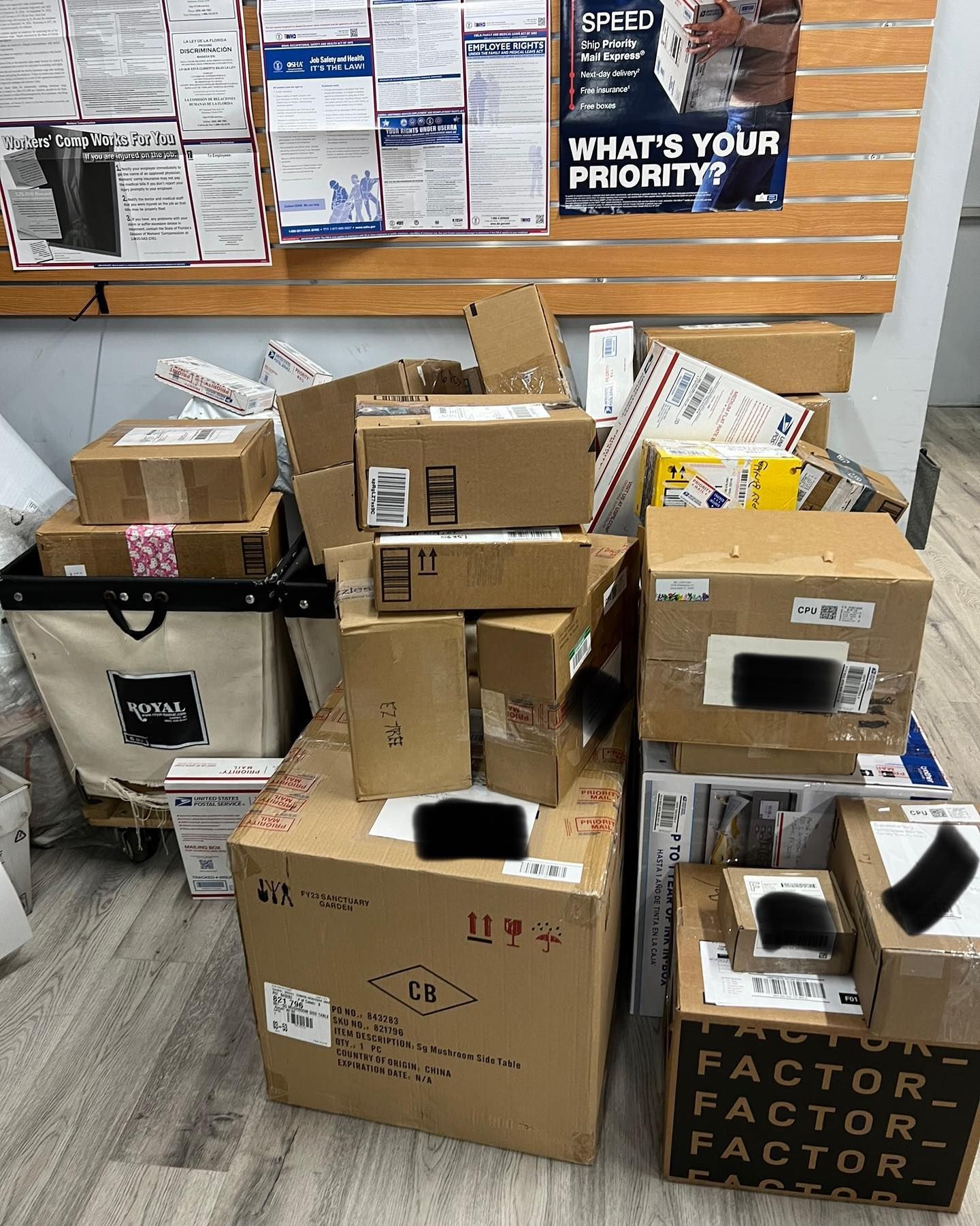 A stack of various cardboard shipping boxes piled in front of a bulletin board on an office wall.