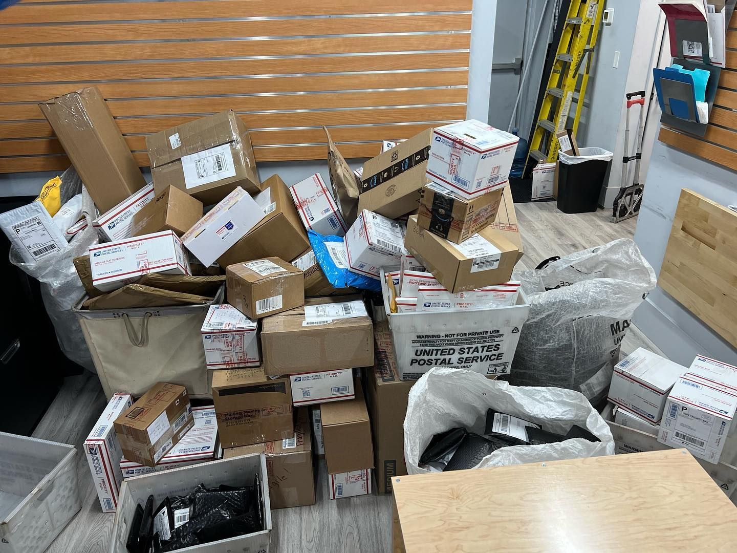 A large pile of various cardboard shipping boxes and mail parcels stacked in an office or postal workspace.