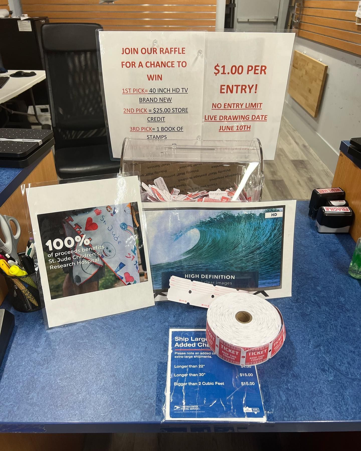 A raffle setup on a blue counter with a prize sign, a clear ticket bin, a roll of raffle tickets, and promotional photos.