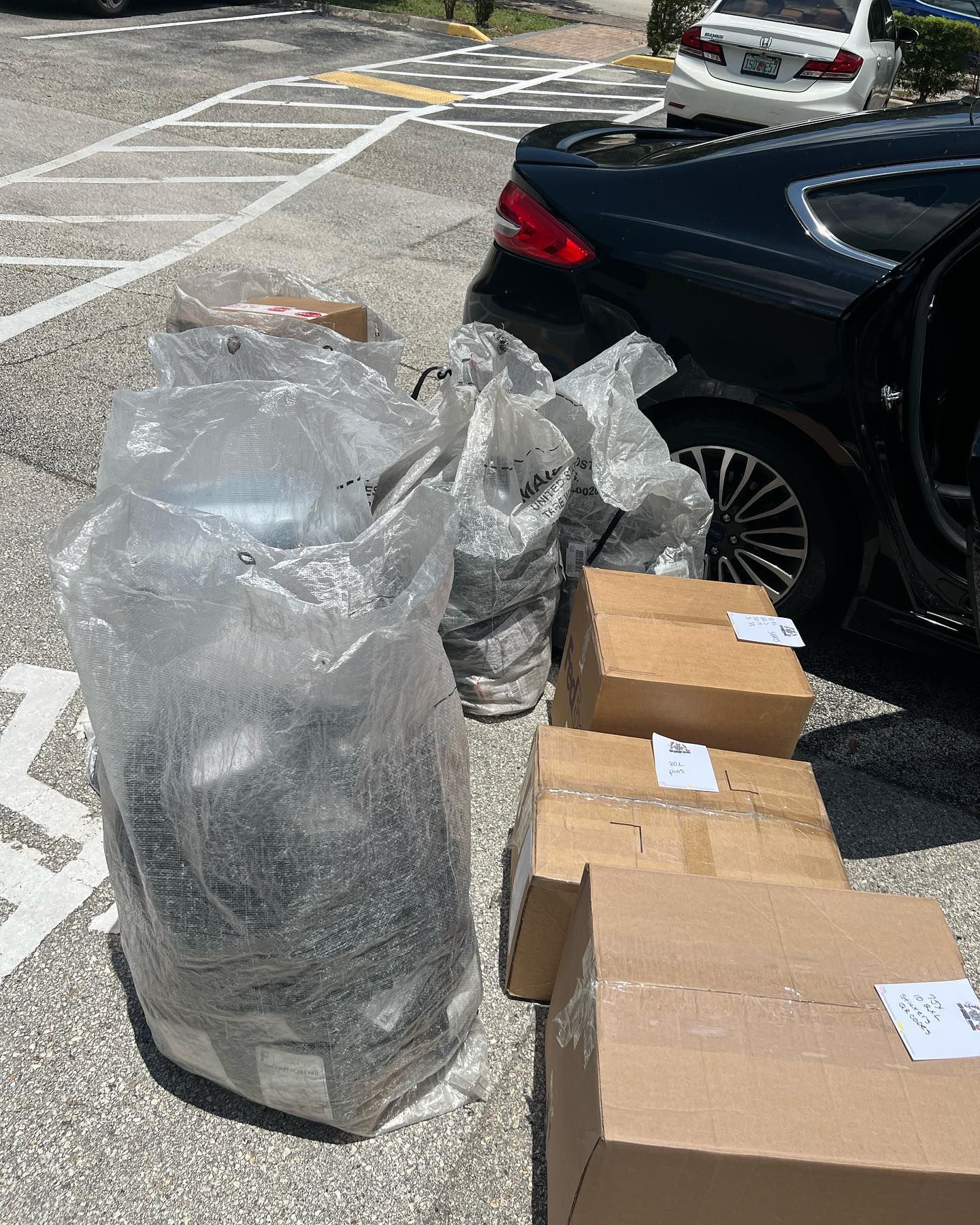Several cardboard boxes and large plastic bags filled with items sit on the asphalt next to a parked black car.