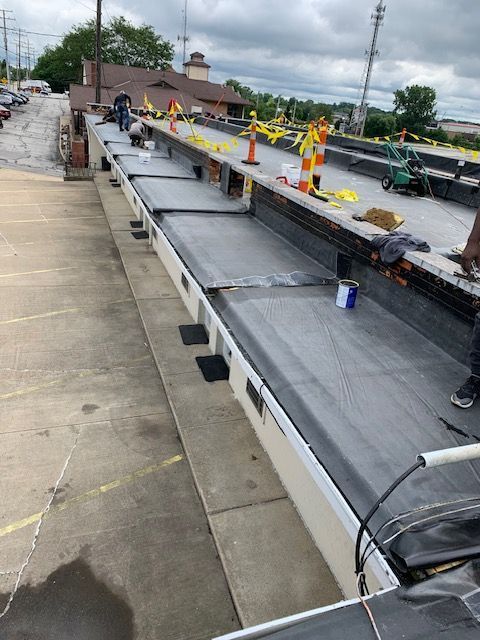 Workers repairing a flat commercial roof, laying black roofing material. Safety cones and caution tape are present.
