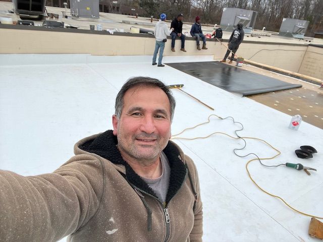 Man takes a selfie on a flat roof with workers in the background; white roofing material and tools are present.