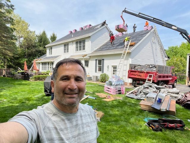 Man taking a selfie in front of a house being re-roofed. Construction workers on roof with crane lifting materials.