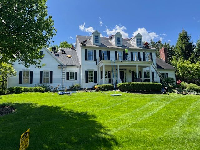 Roofers working on a large white house with black shutters and a green lawn on a sunny day.