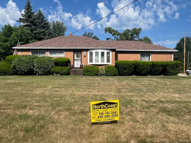 Ranch-style house with brown roof and brick exterior, surrounded by green shrubs and a grassy yard. A sign is in the foreground.