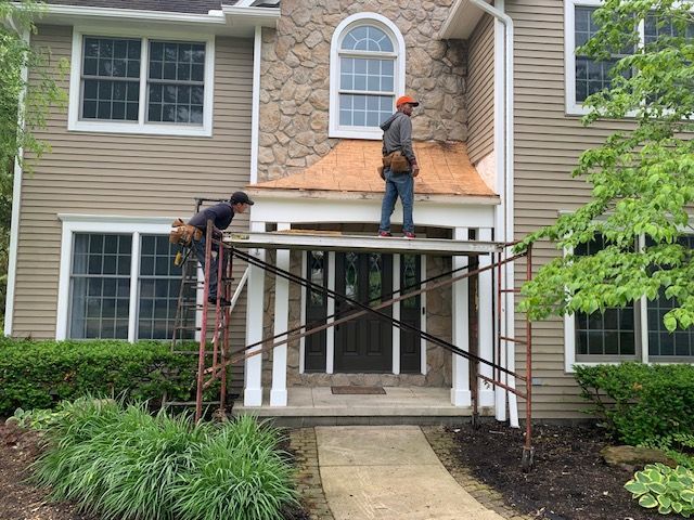 Two workers on scaffolding repair a house's entryway roof. Beige siding, stone facade, and green bushes.