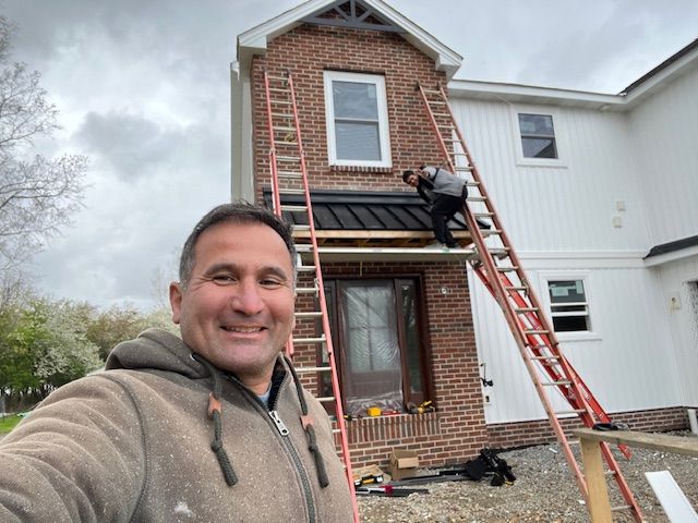 Man taking selfie in front of a house under construction. Brick and white siding. Two workers on ladders.