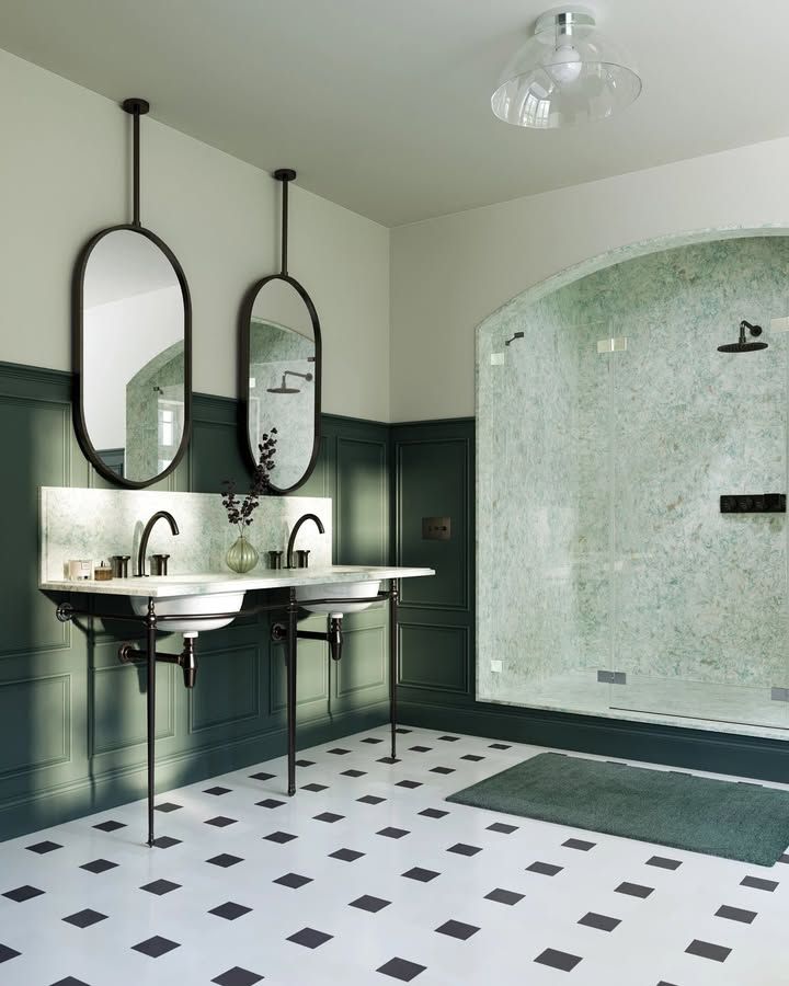 Bathroom with white tile floor, dark green wainscoting, double vanity with oval mirrors, and a glass-enclosed shower.