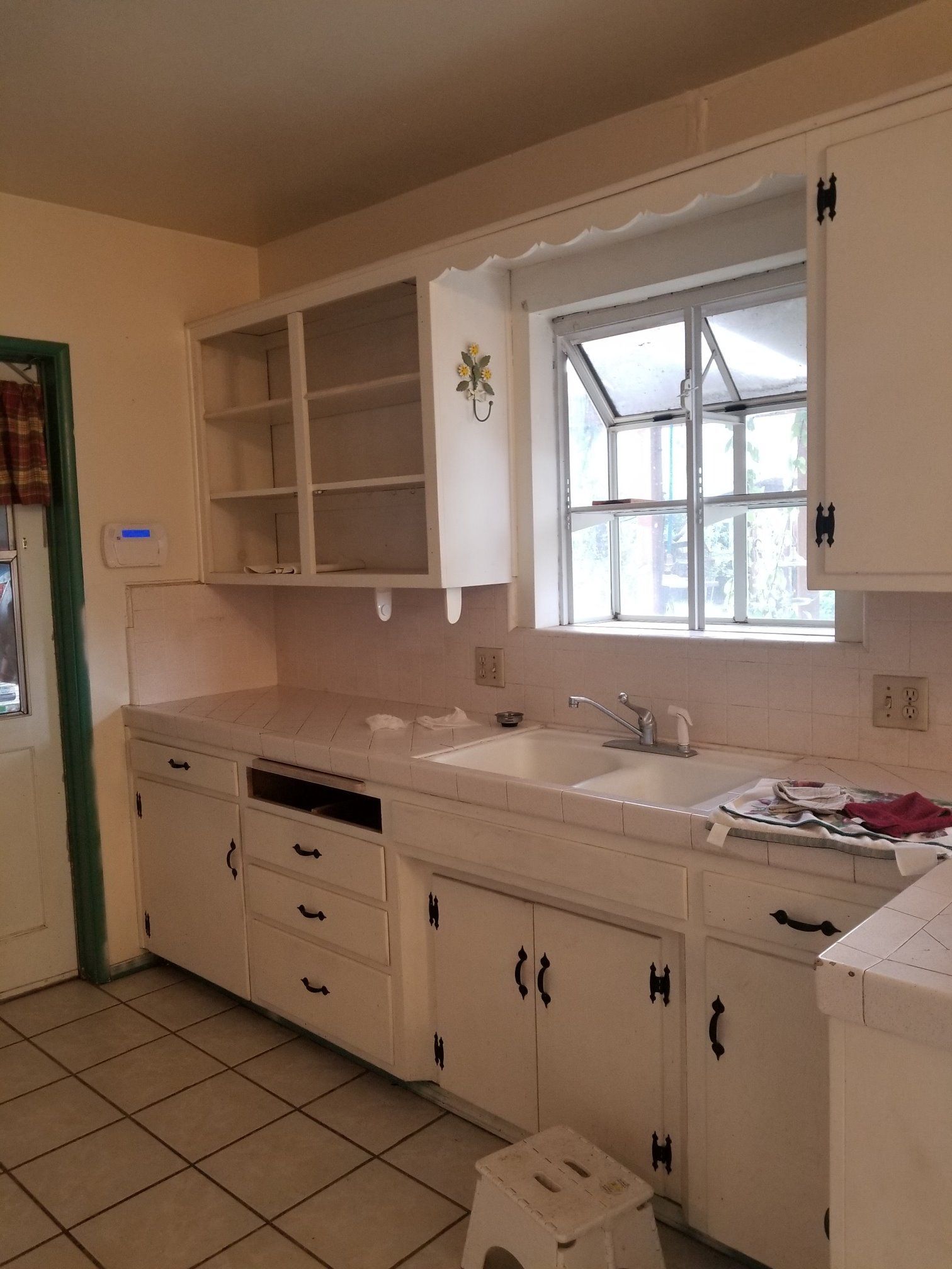 White kitchen cabinets, countertop, and sink. Window above sink. White tile floor. Doorway on left.