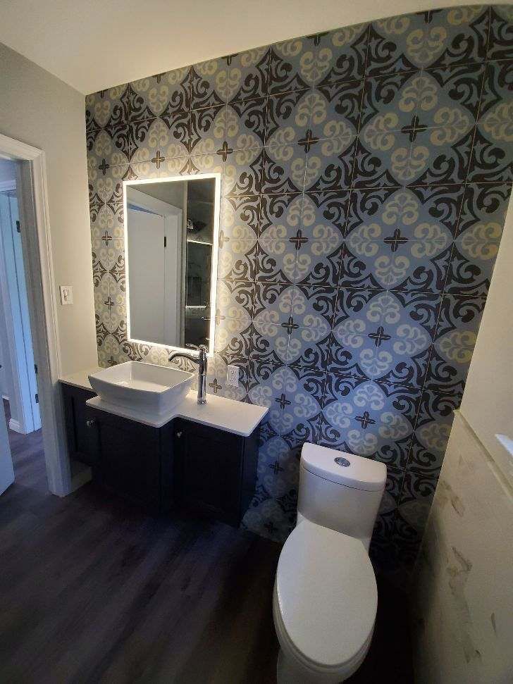 Bathroom with patterned wallpaper, illuminated mirror, dark vanity, white sink and toilet.
