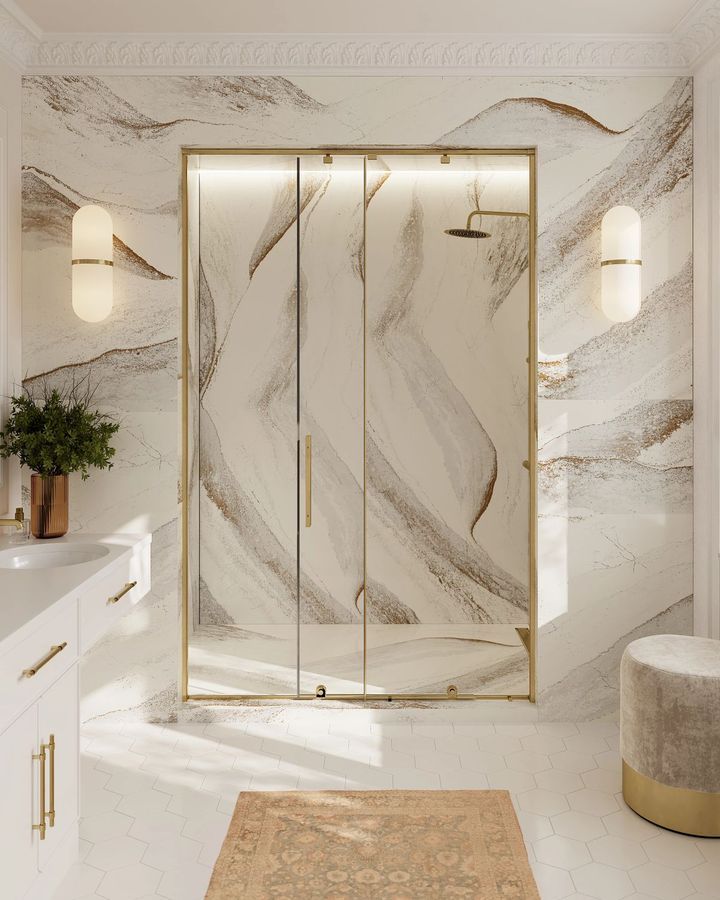 Bathroom with gold-framed glass shower, marble-effect walls, gold accents, and a beige rug.